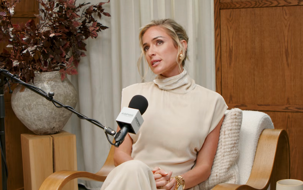 A person in a high-neck, sleeveless outfit is seated, speaking into a microphone, with decorative plants and a textured blanket in the background