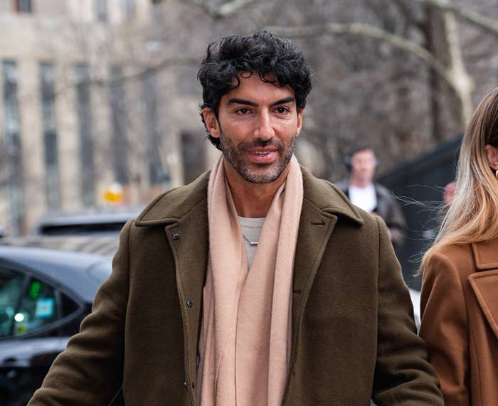 A man with curly hair in a brown coat and beige scarf walking outside, likely in an urban setting. He appears casual and comfortable