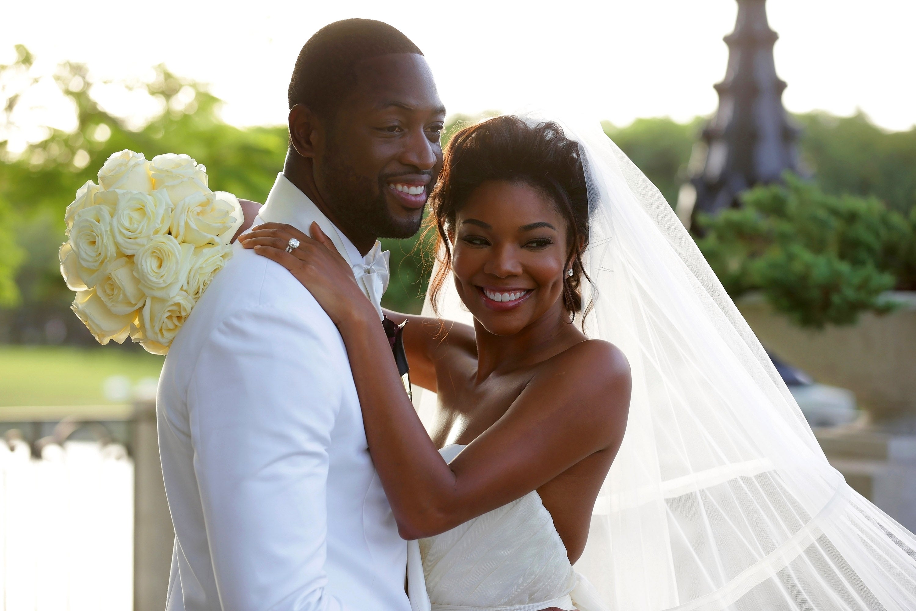 A bride in an elegant strapless gown and veil smiles while embracing a man in a white suit with roses in the background