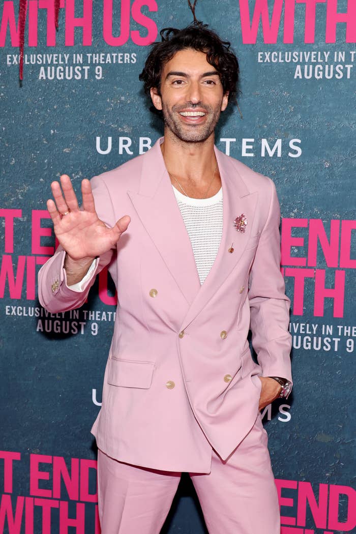 Person in a stylish pink suit with a white shirt waves at a "It Ends With Us" event backdrop