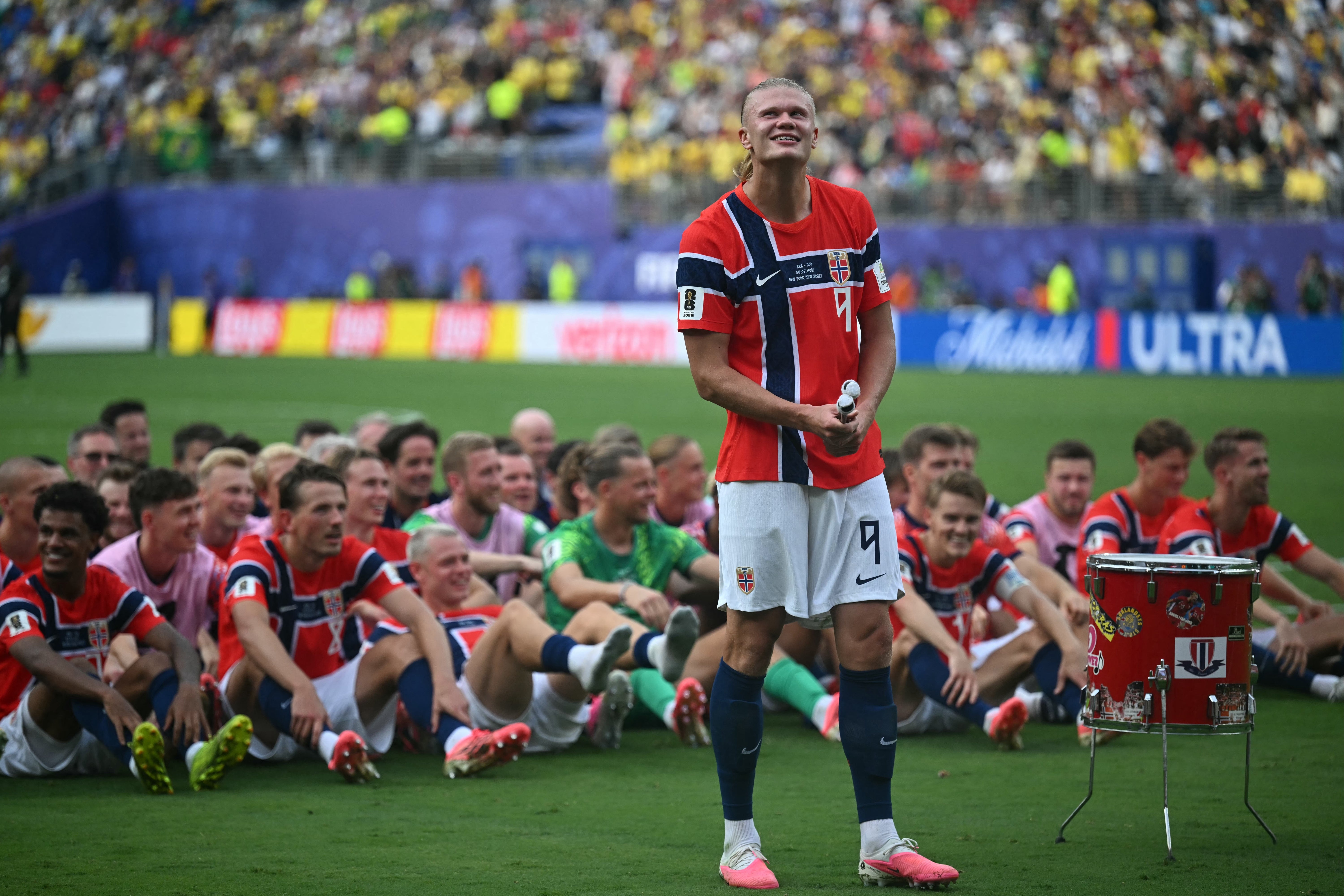 Fotballspiller på en bane, smilende og holder rullet papir, med lagkamerater som sitter bak ham. Stadionpublikum i bakgrunnen