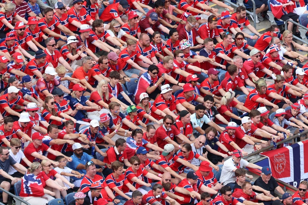 Fans i matchende antrekk utfører en synkronisert gest på et stadion, og viser samhold og entusiasme. Norsk flagg synlig