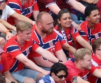 Fans i matchende antrekk utfører en synkronisert gest på et stadion, og viser samhold og entusiasme. Norsk flagg synlig