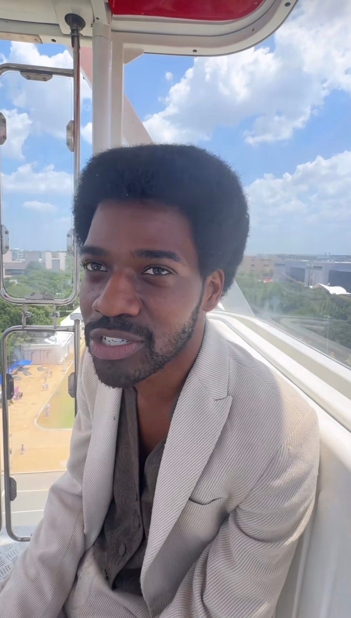 Mann med afro-frisyre i lys dress sitter i en pariserhjulshytte med utsikt over bybildet utenfor