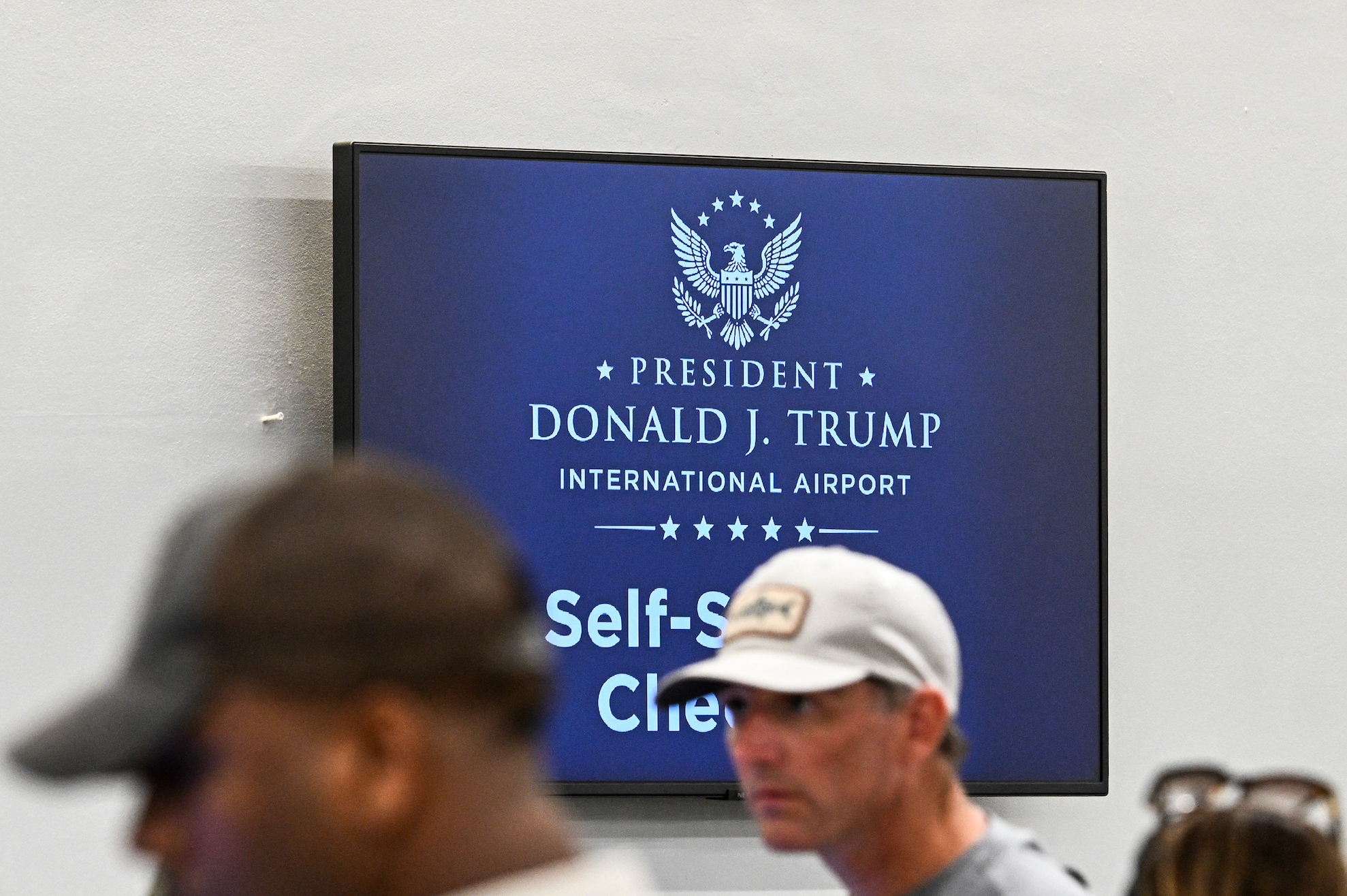 Skiltet på flyplassen viser "President Donald J. Trump International Airport, Self-Service Check-In" med mennesker uskarpe i forgrunnen