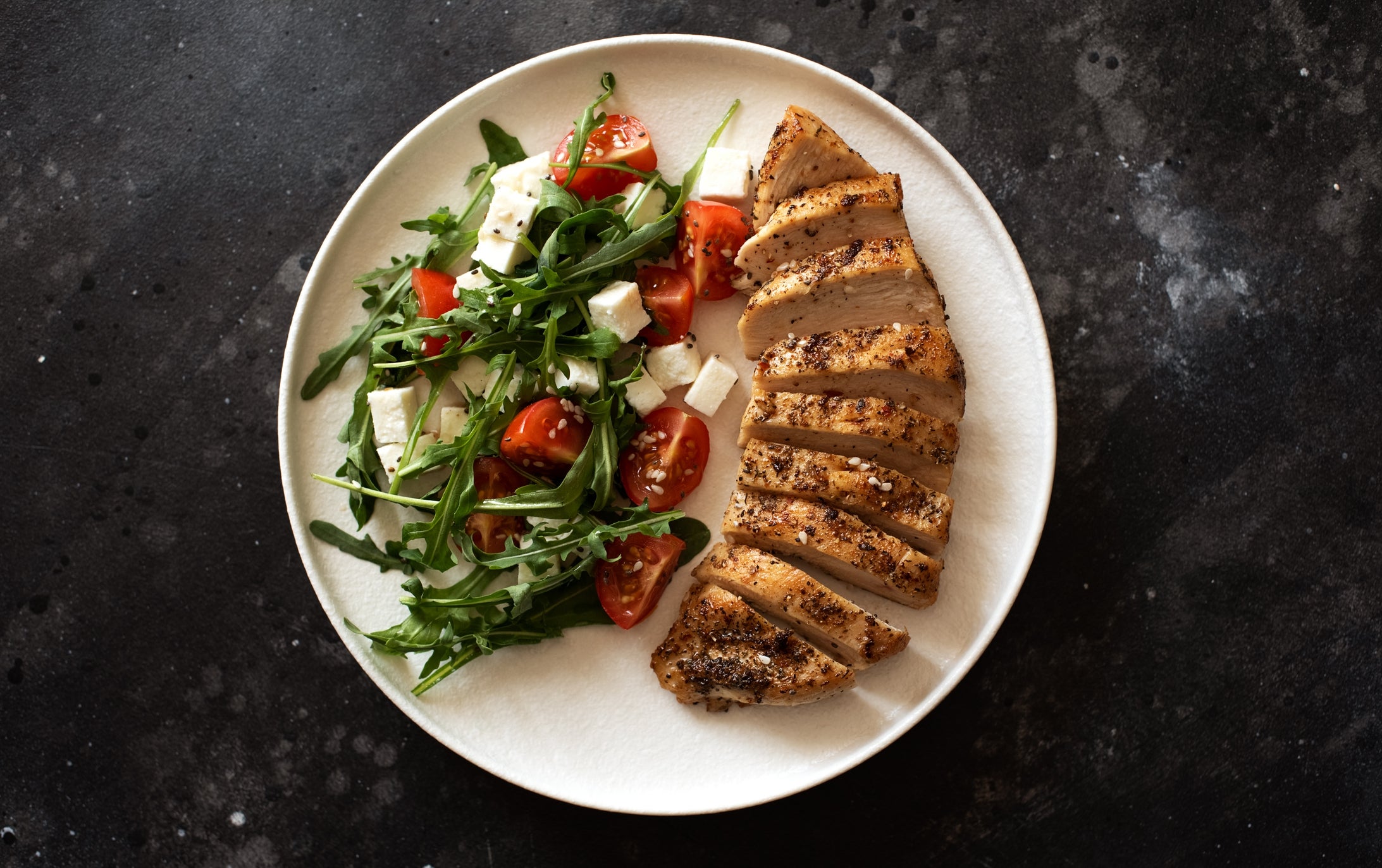 Grilled chicken slices with a side salad of arugula, cherry tomatoes, and feta cheese on a round plate
