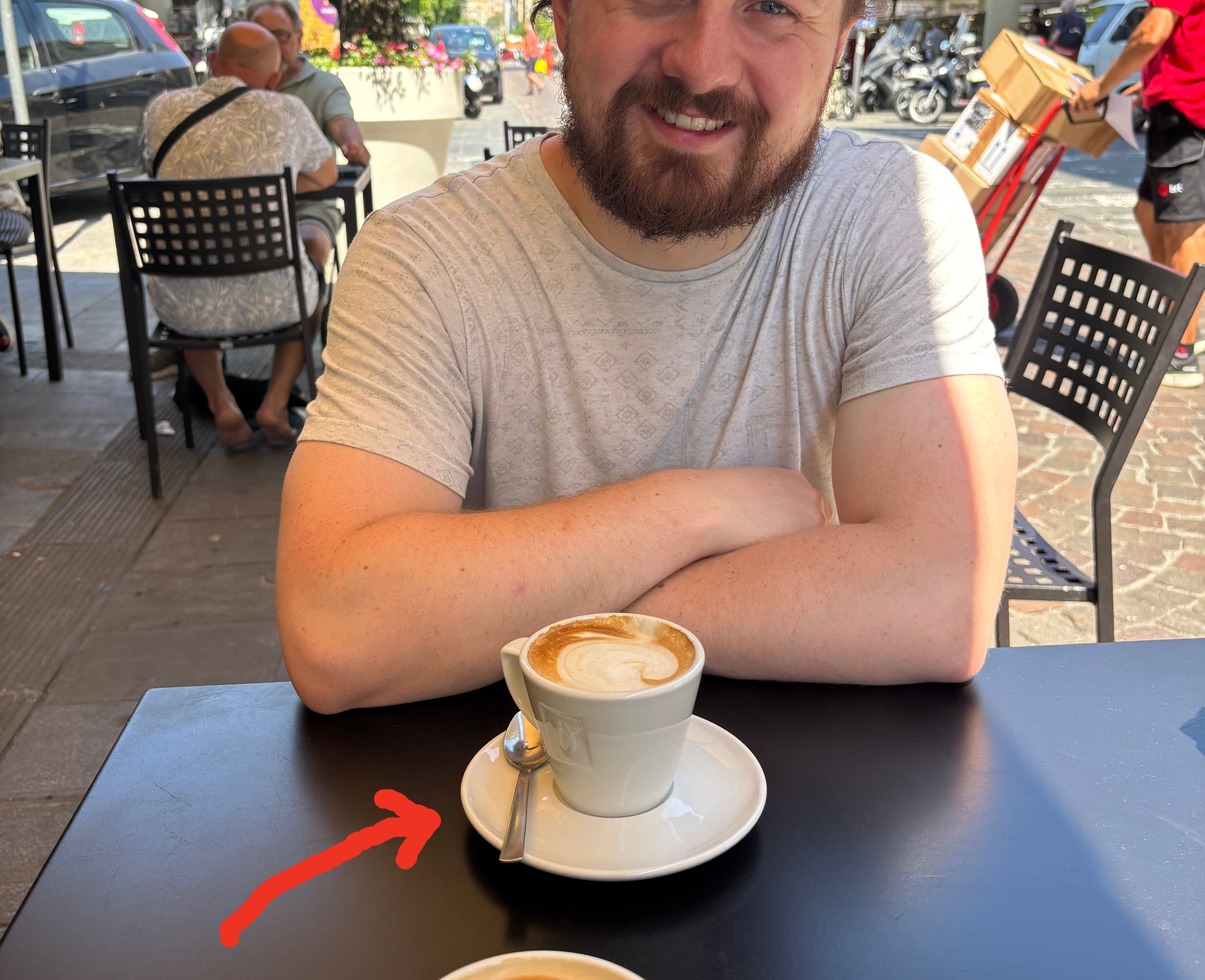 A person seated at an outdoor cafe table with two cups of coffee in front. Background shows a street scene with pedestrians