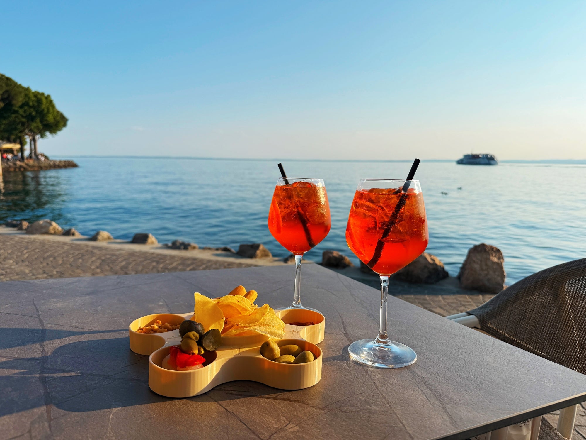 Two Aperol spritz cocktails on a table by a calm seaside, with snacks in a small tray, evoking a relaxing travel vibe