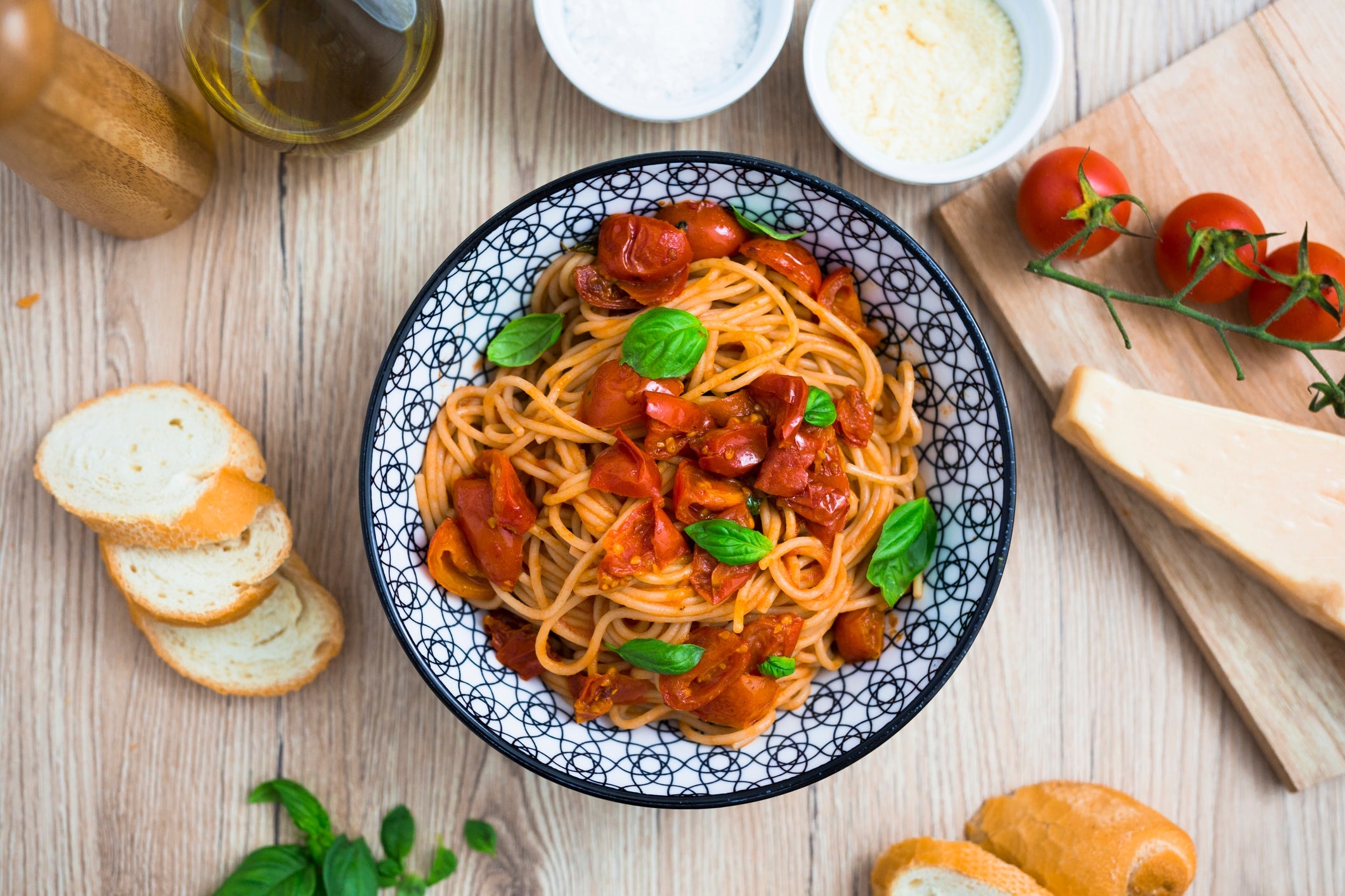 A bowl of pasta topped with tomatoes and basil sits on a wooden table, surrounded by bread, cherry tomatoes, and cheese