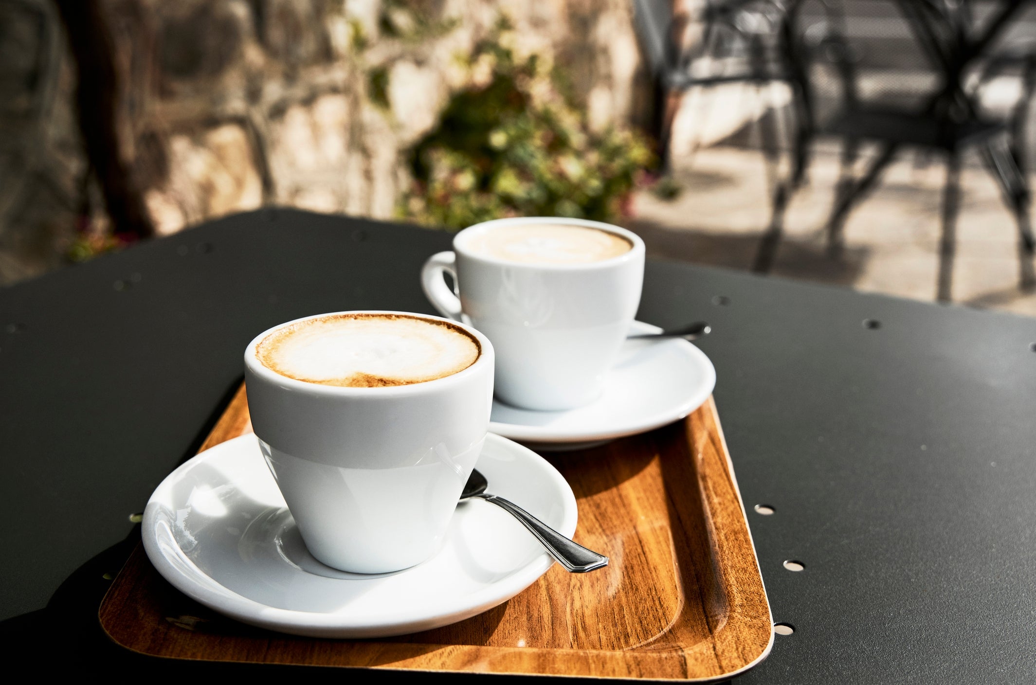Two cups of coffee on a wooden tray, placed on an outdoor table at a café, with blurred seating area in the background
