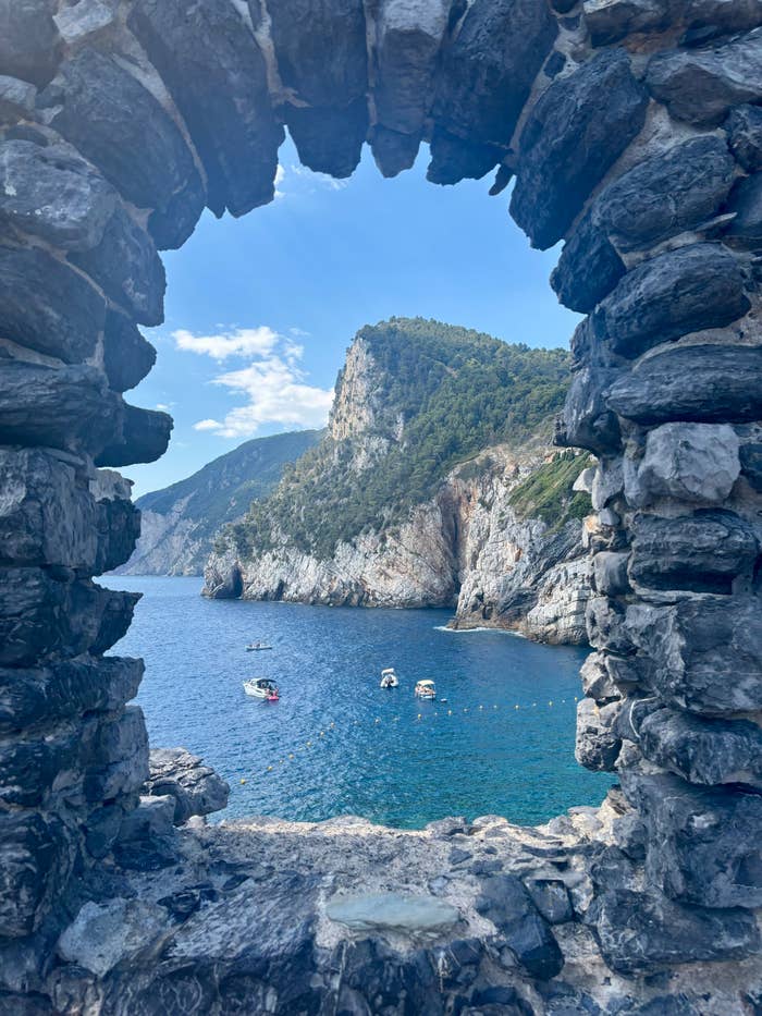 View through a stone arch of a rocky coastline with several small boats on a blue sea