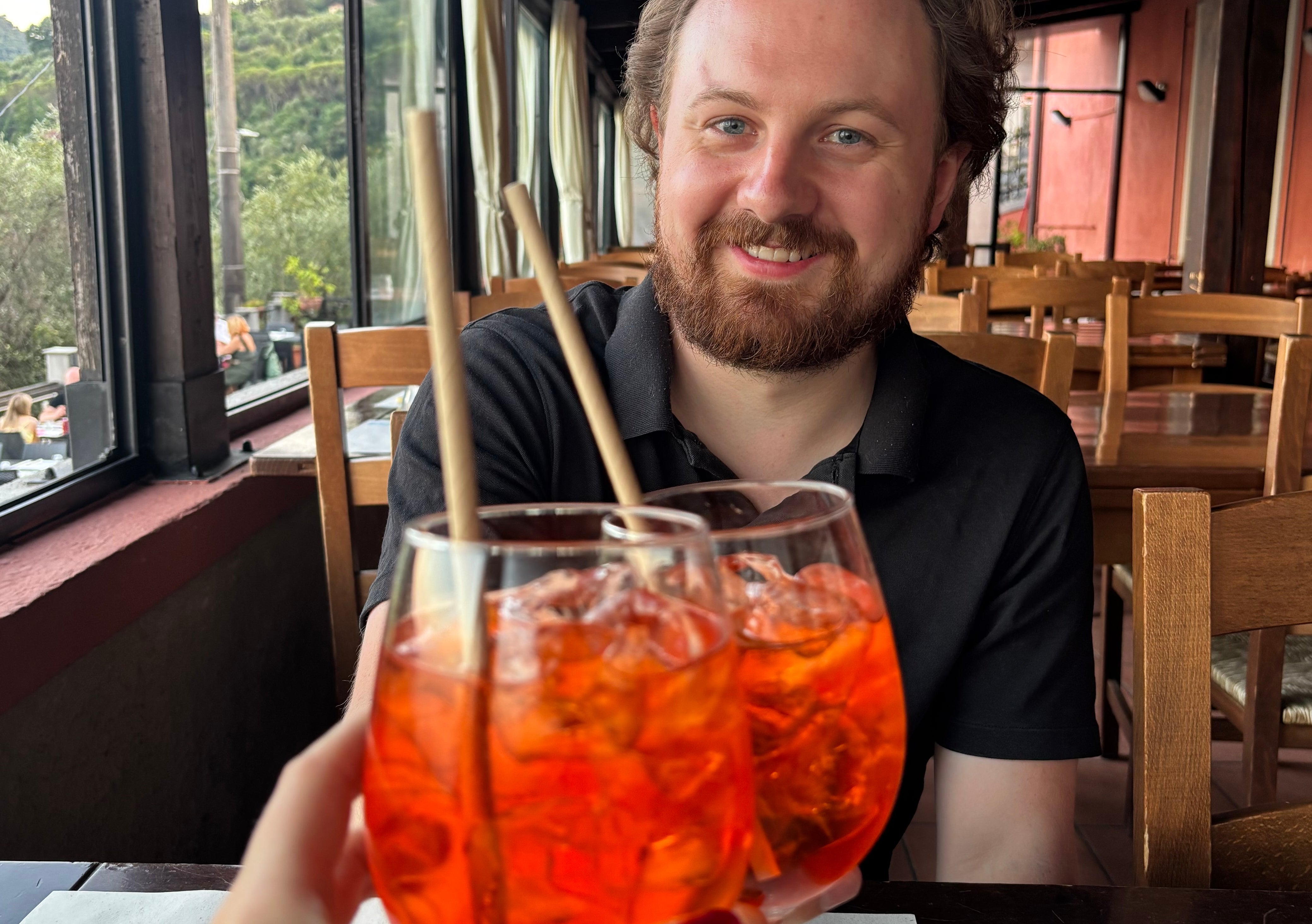 A person sits at a restaurant table, smiling across from two glasses of iced drinks raised in a toast, with an outdoor view in the background