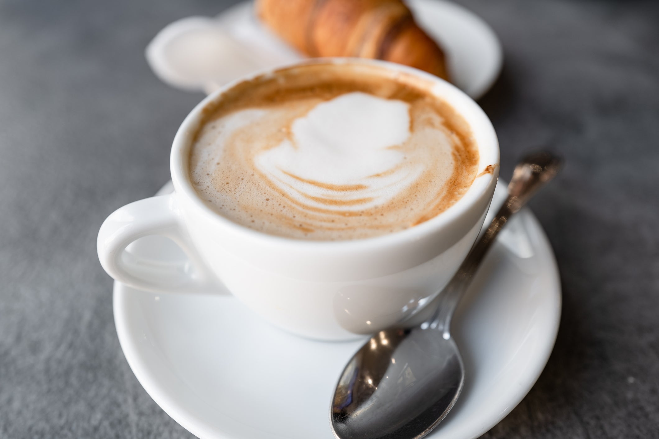 A cup of cappuccino with frothy milk art, served on a saucer with a spoon, with a croissant in the background