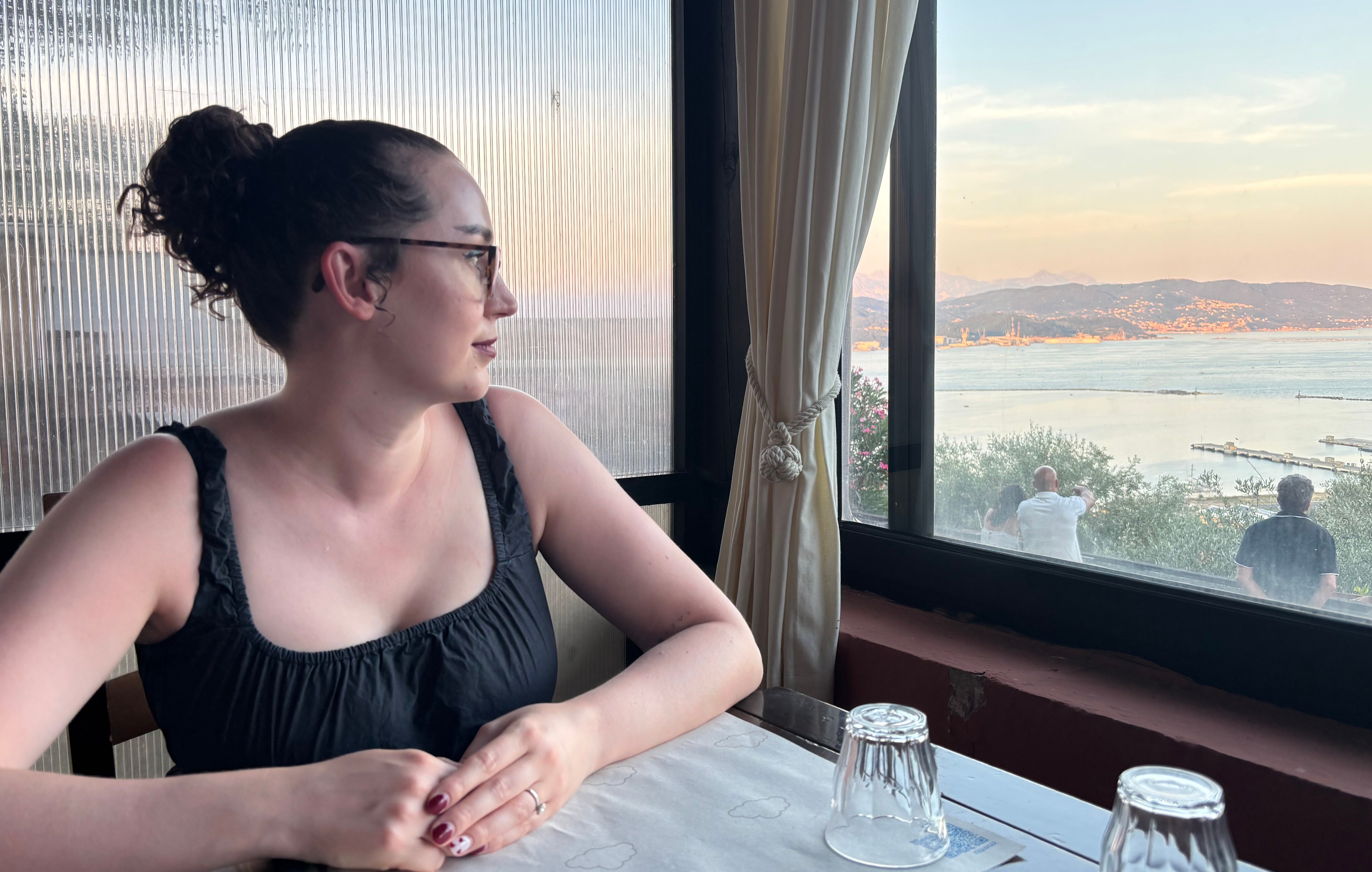 Person seated at a table by a large window, gazing at a scenic coastal view with water and distant hills in the background