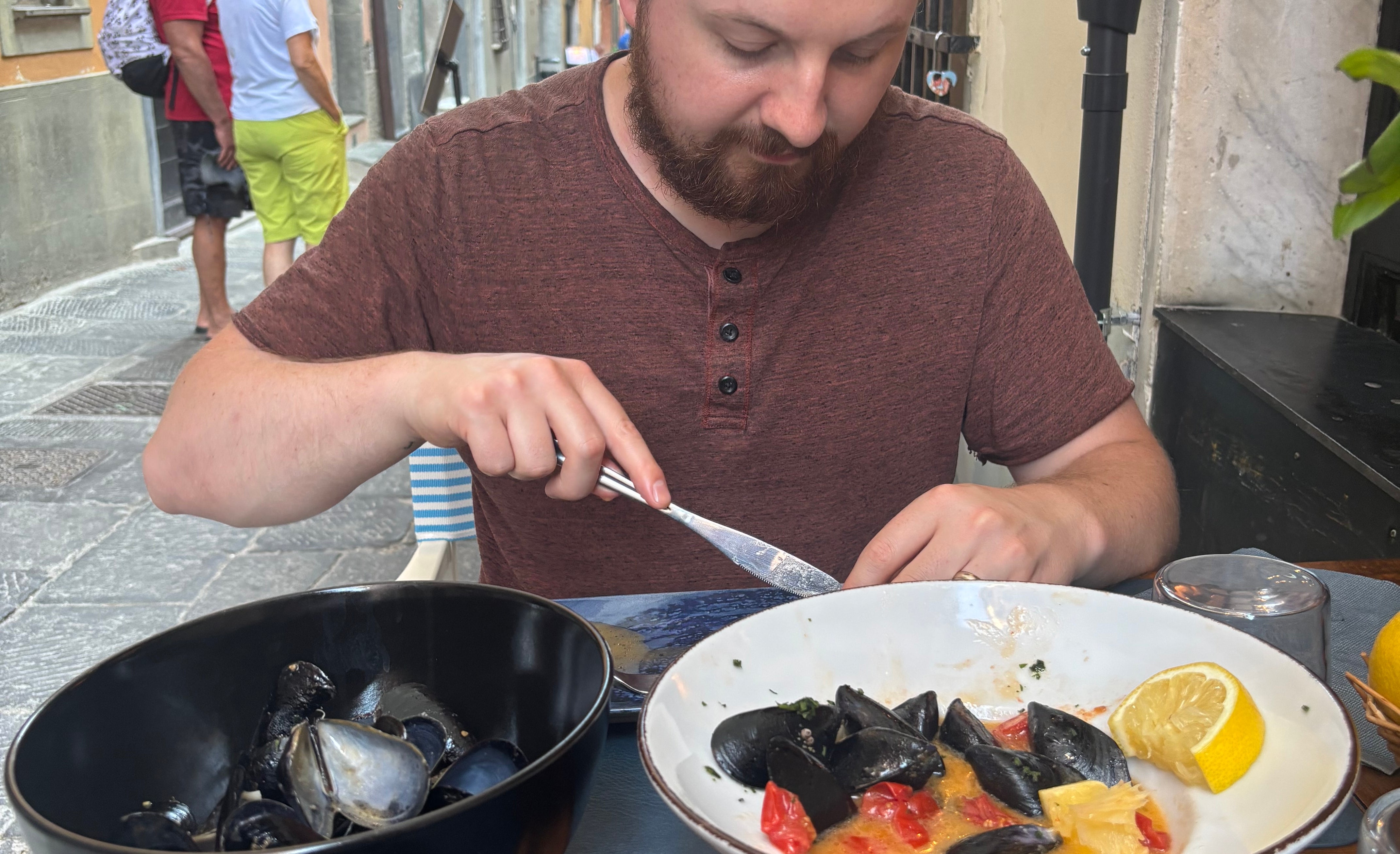 Person dining outdoors, enjoying a seafood dish at a quaint European street café. Other diners are visible in the background