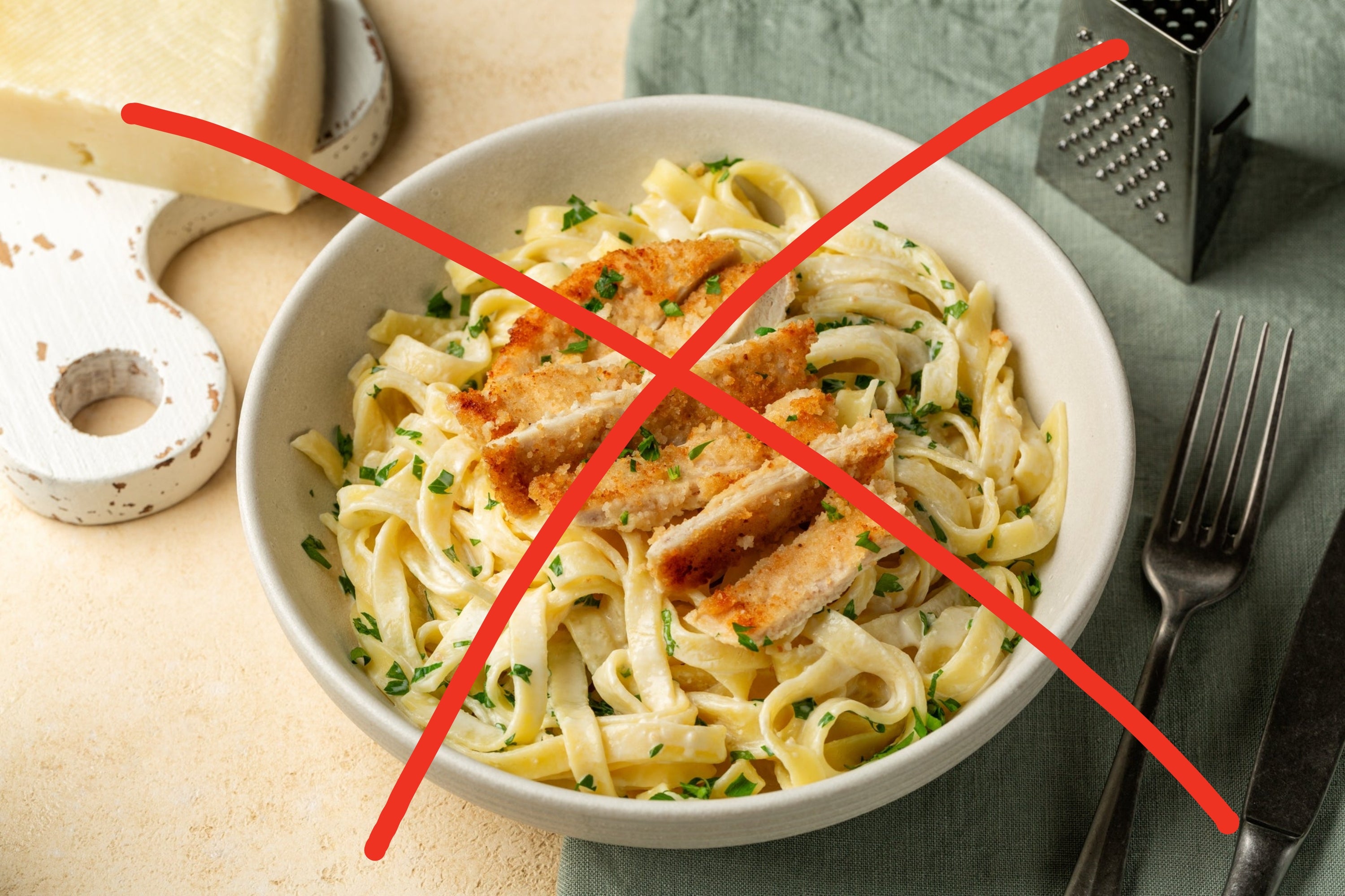 A bowl of fettuccine Alfredo topped with sliced breaded chicken, garnished with herbs. Cheese and grater in the background