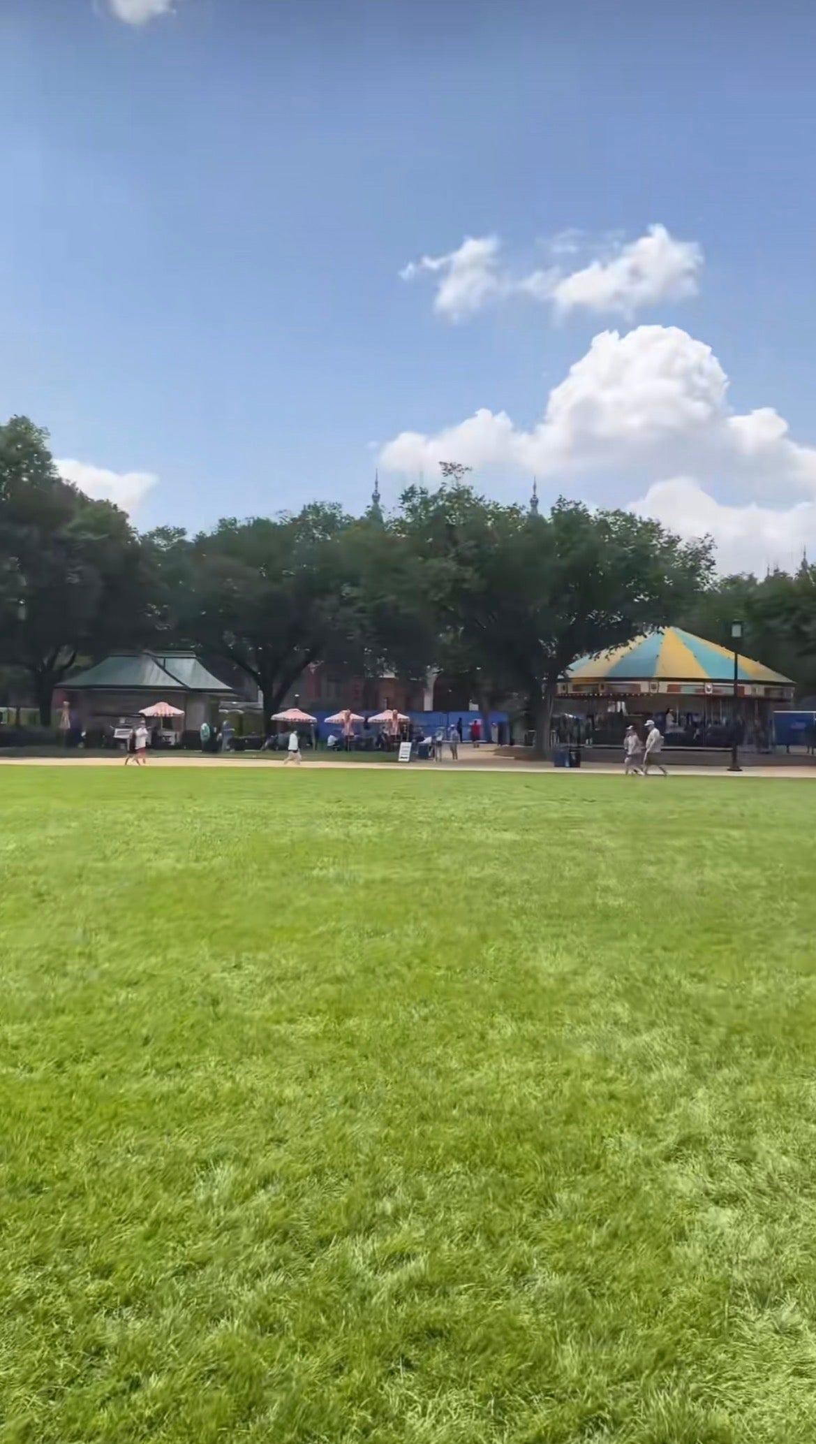 Åpen parkscene med folk som går nær fargerike telt og trær i bakgrunnen. Lys, klar himmel med luftige skyer