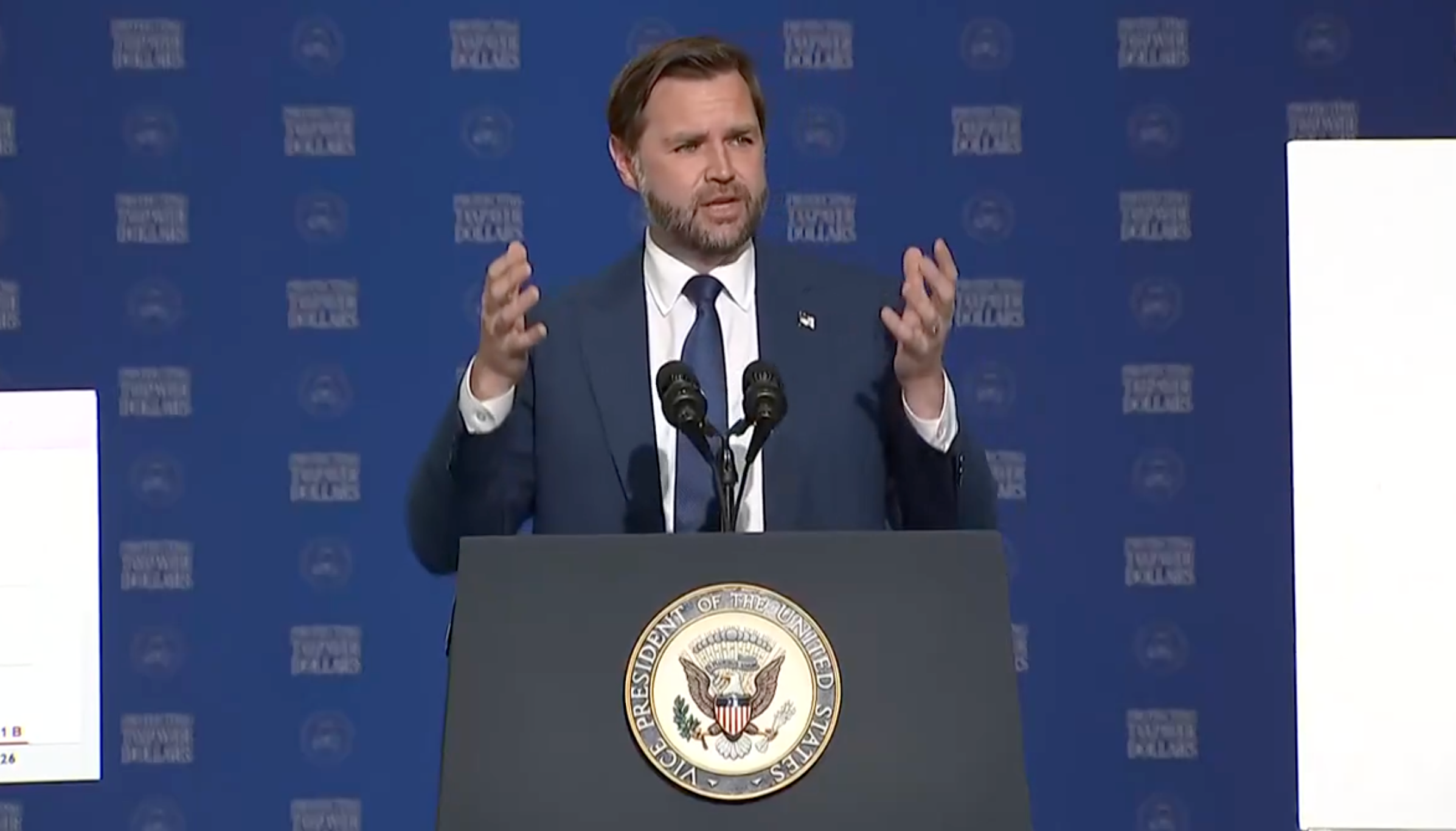 En mann i dress snakker ved et podium med et amerikansk segl og gestikulerer med hendene. Bakgrunnen viser logoer for sponsing av arrangementer