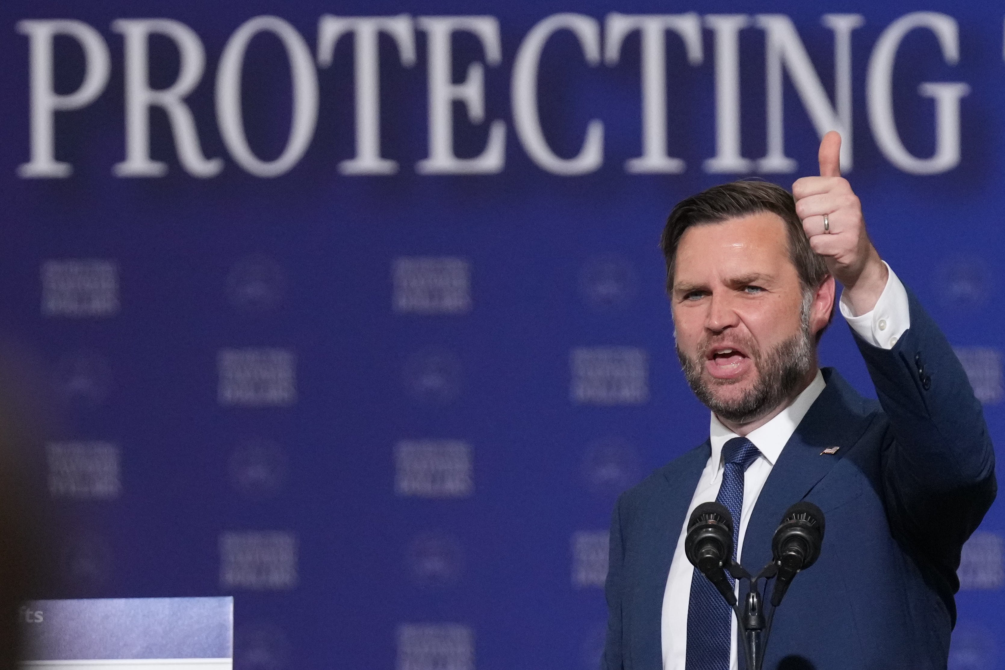 Man successful a suit giving a thumbs-up motion portion speaking astatine a podium, with ample "PROTECTING" substance down him