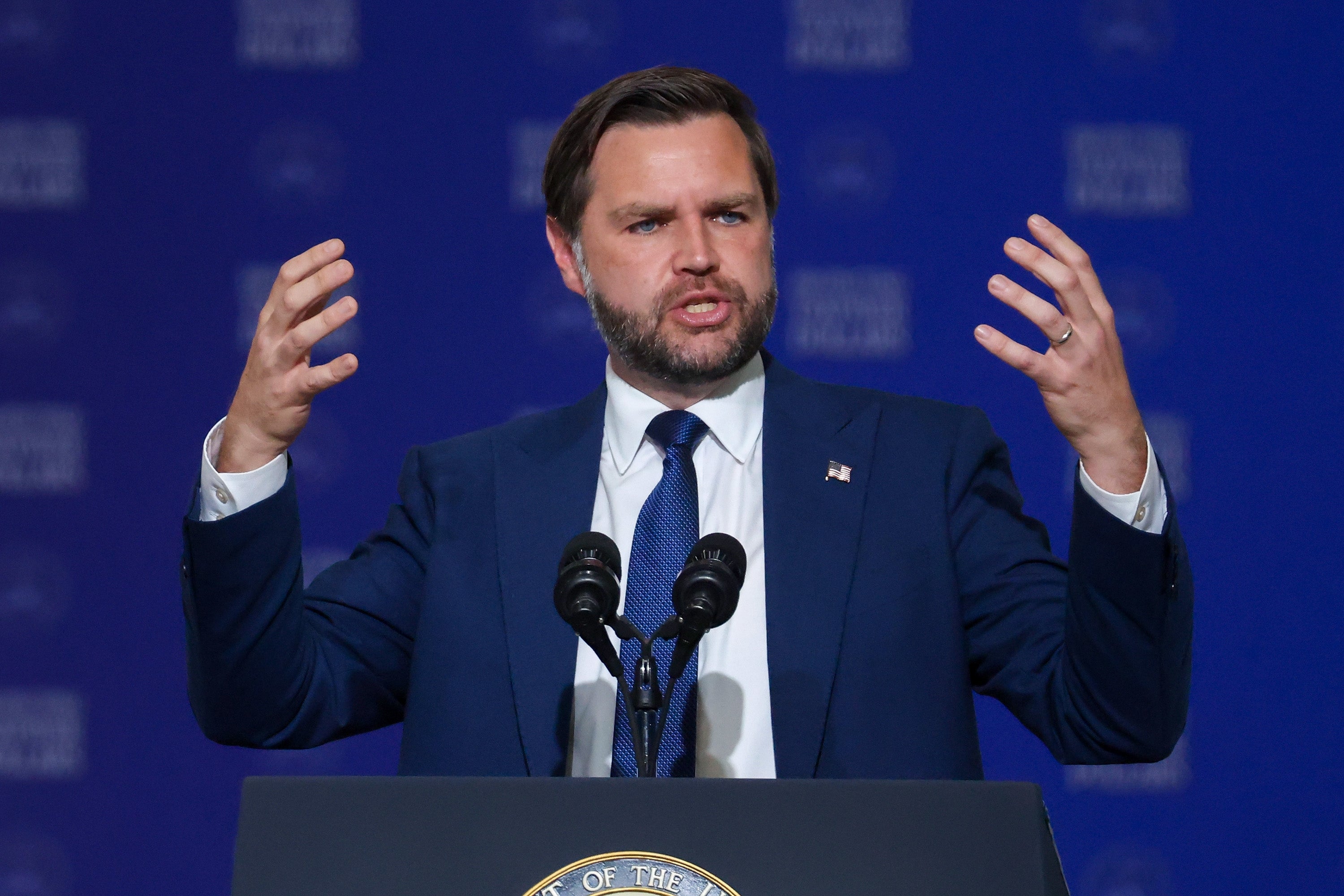 Person astatine a podium speaking, gesturing with raised hands, dressed successful a suit and tie, with a bluish backdrop
