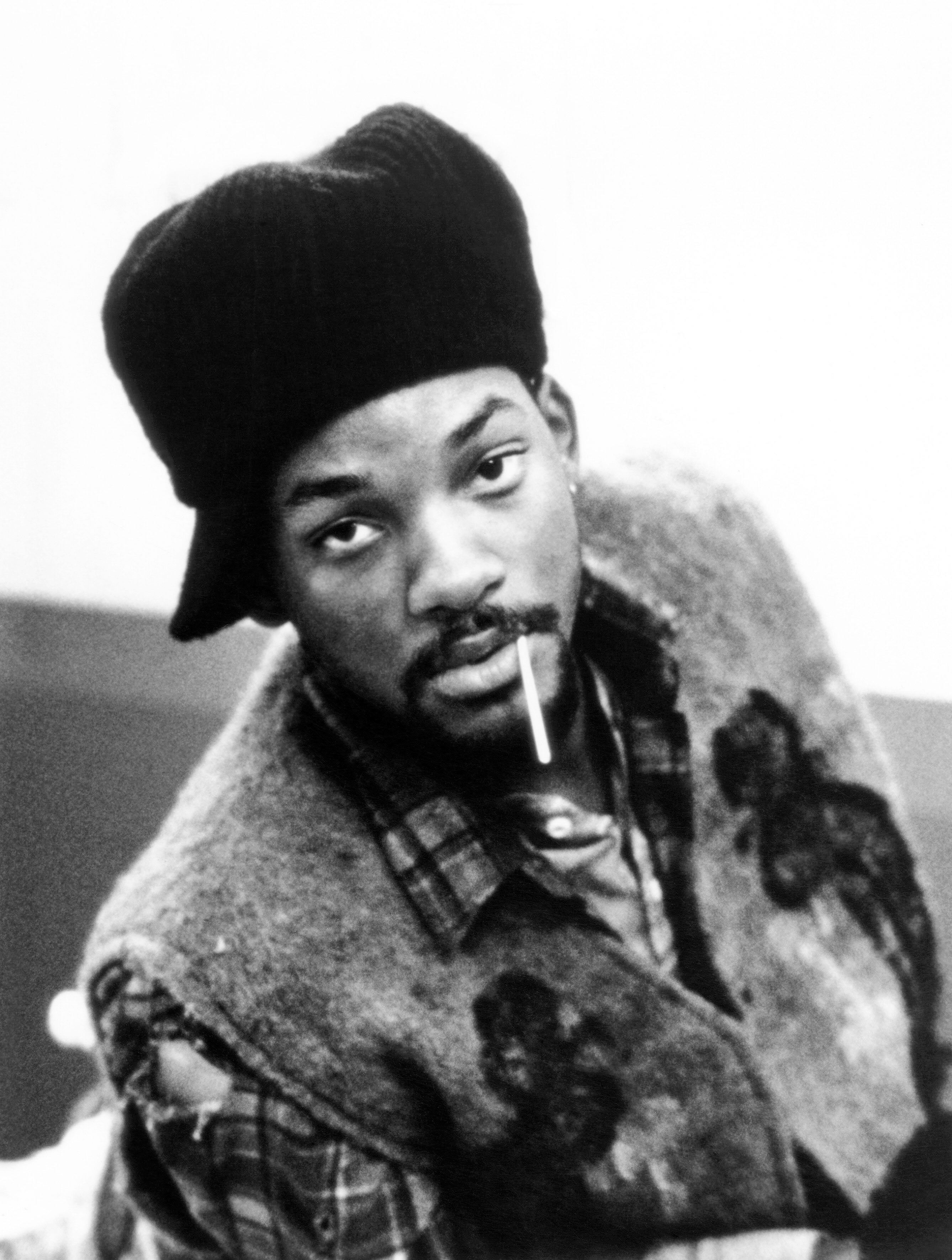 Younger Will Smith wearing a knit hat and patterned vest, holding a toothpick in their mouth, gives a relaxed and contemplative look