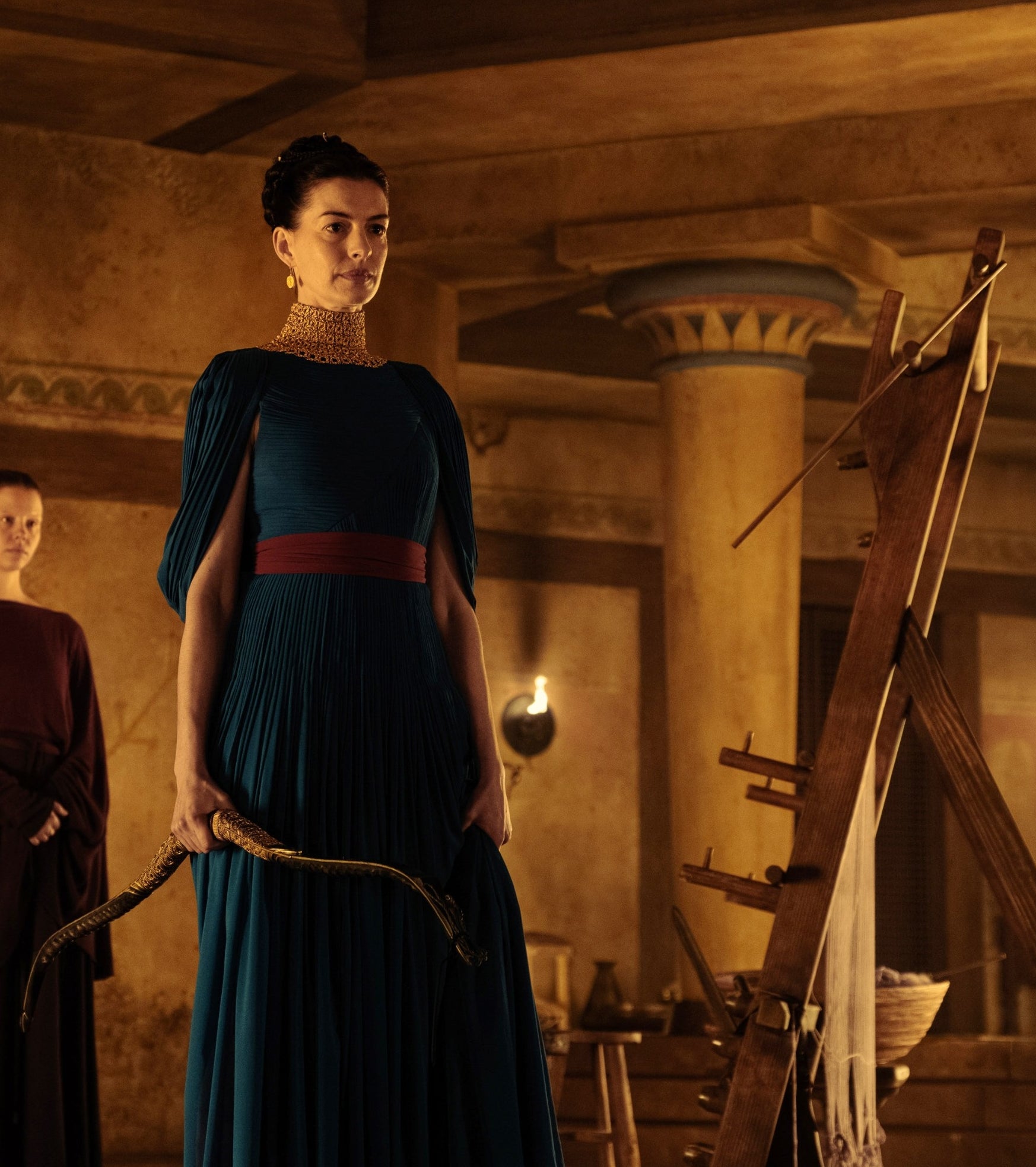 Anne Hathaway in historical costume stands indoors with a bow, in a room with ancient decor