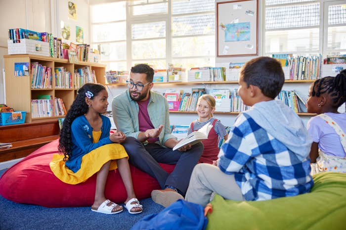 A teacher sits with a group of children in a library, engaging them during a reading session. Books are visible on shelves in the background