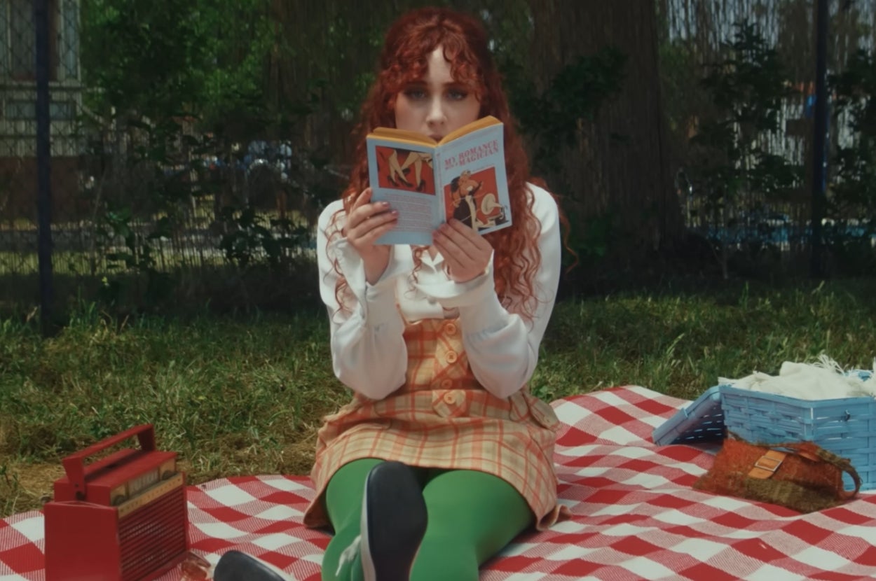 Chappel Roan seated on a picnic blanket, reading a book. They have long curly hair and wear a layered outfit with green tights