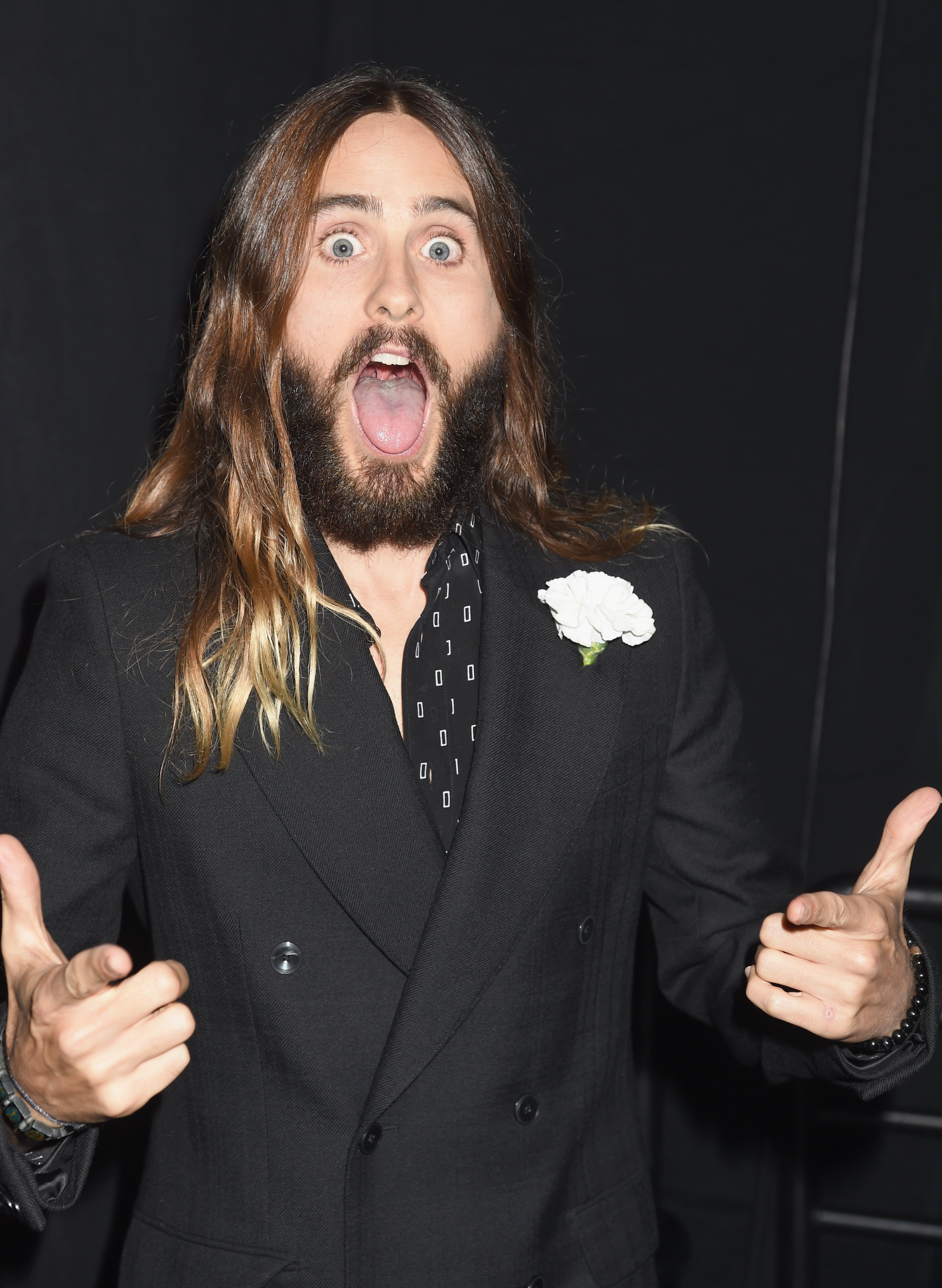Jared Leto, with long hair and a beard, dressed in a double-breasted black suit, energetically gesturing with both hands