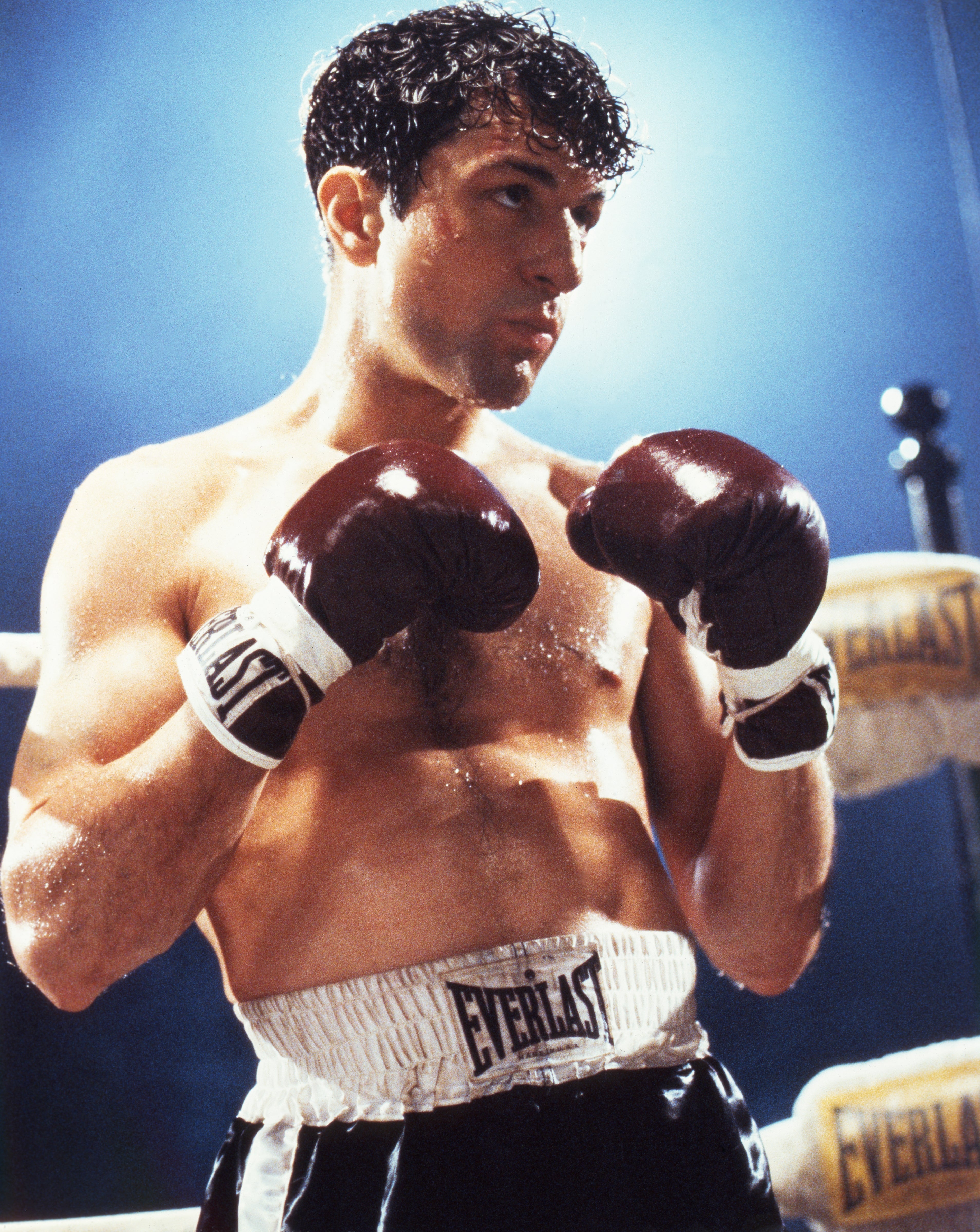 Robert De Niro portrays the boxer, Jake LaMotta. He wears boxing gloves and shorts while pensively gazing to the side