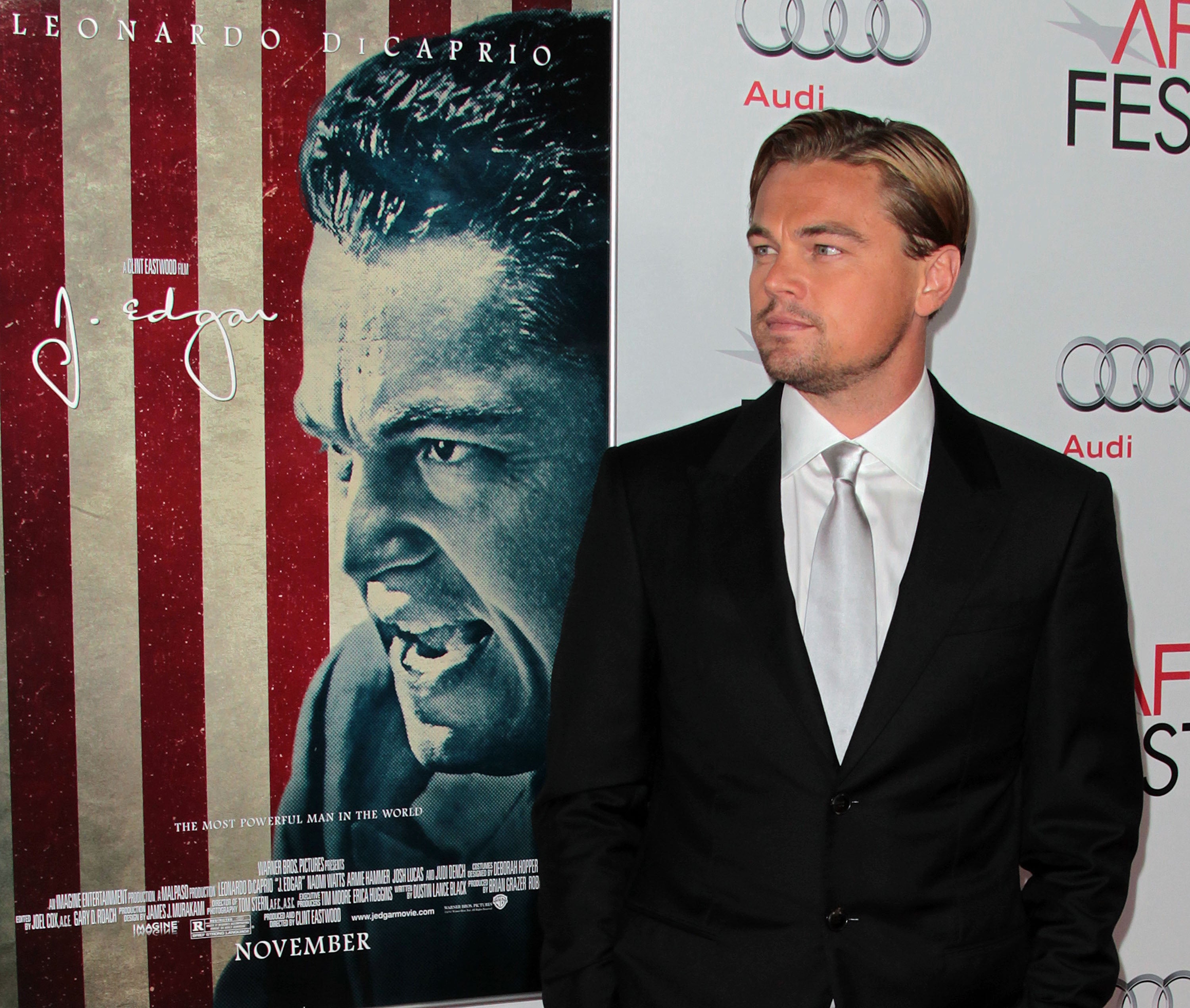 Leonardo DiCaprio stands beside a movie poster for J. Edgar