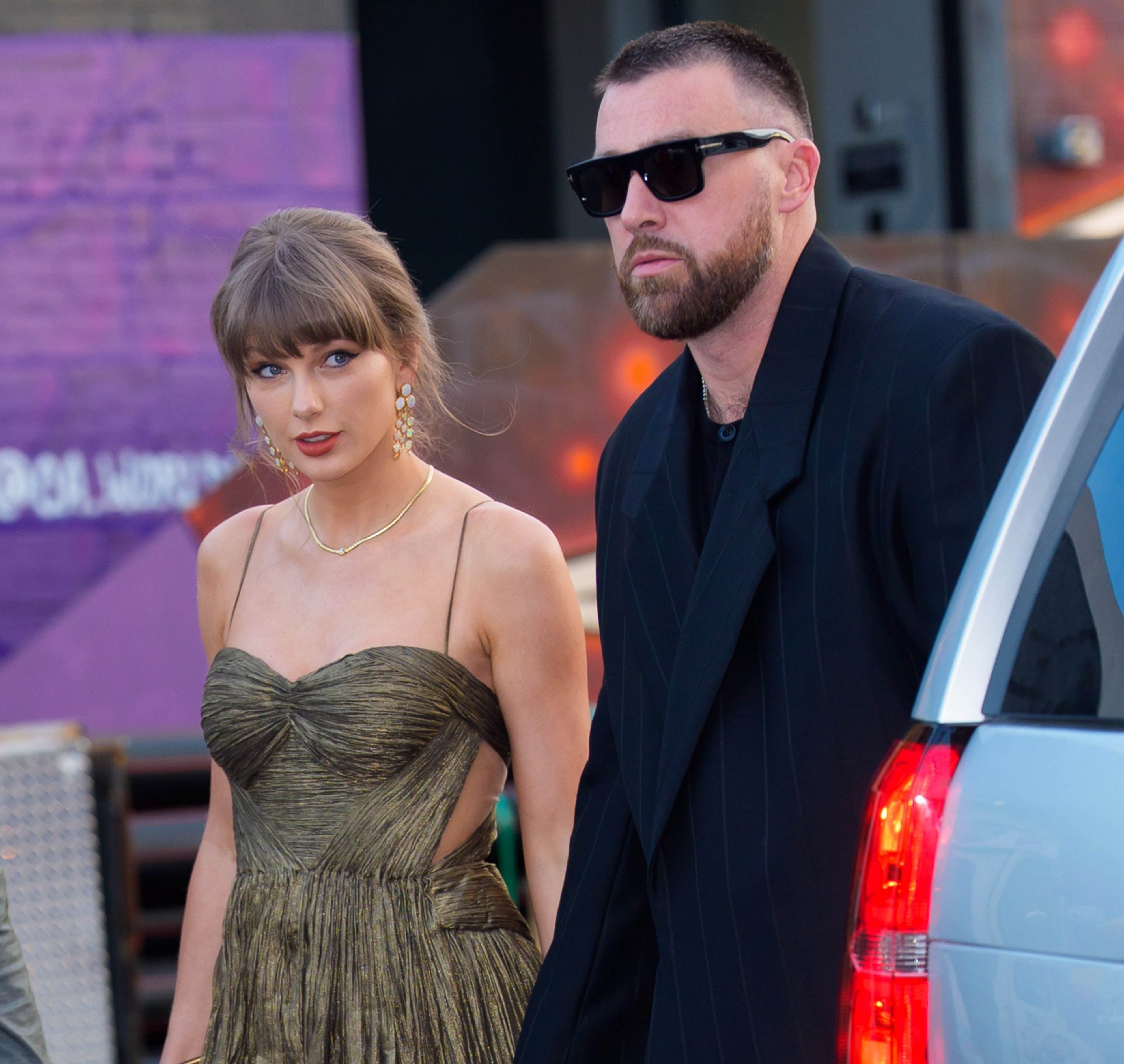 A person in an elegant, strapless dress holds hands with a person in a dark suit and sunglasses on a public street