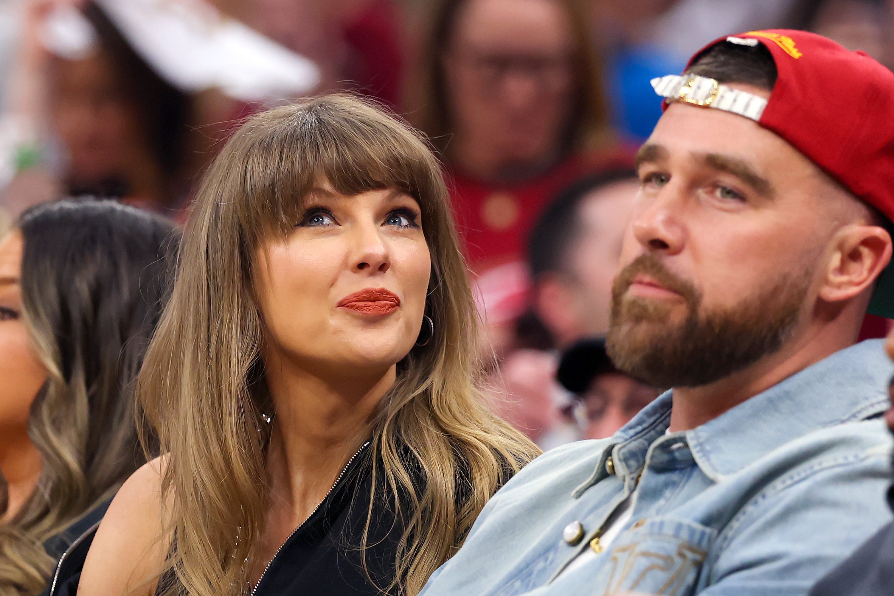 A woman in elegant attire sits beside a man in casual wear and a cap at a public event, both looking forward