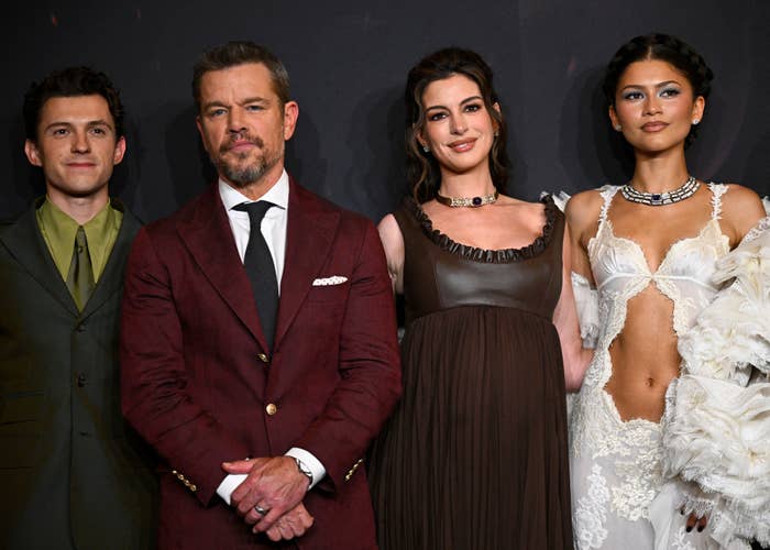 Four people on a red carpet; one in a suit, and three in stylish outfits featuring intricate designs