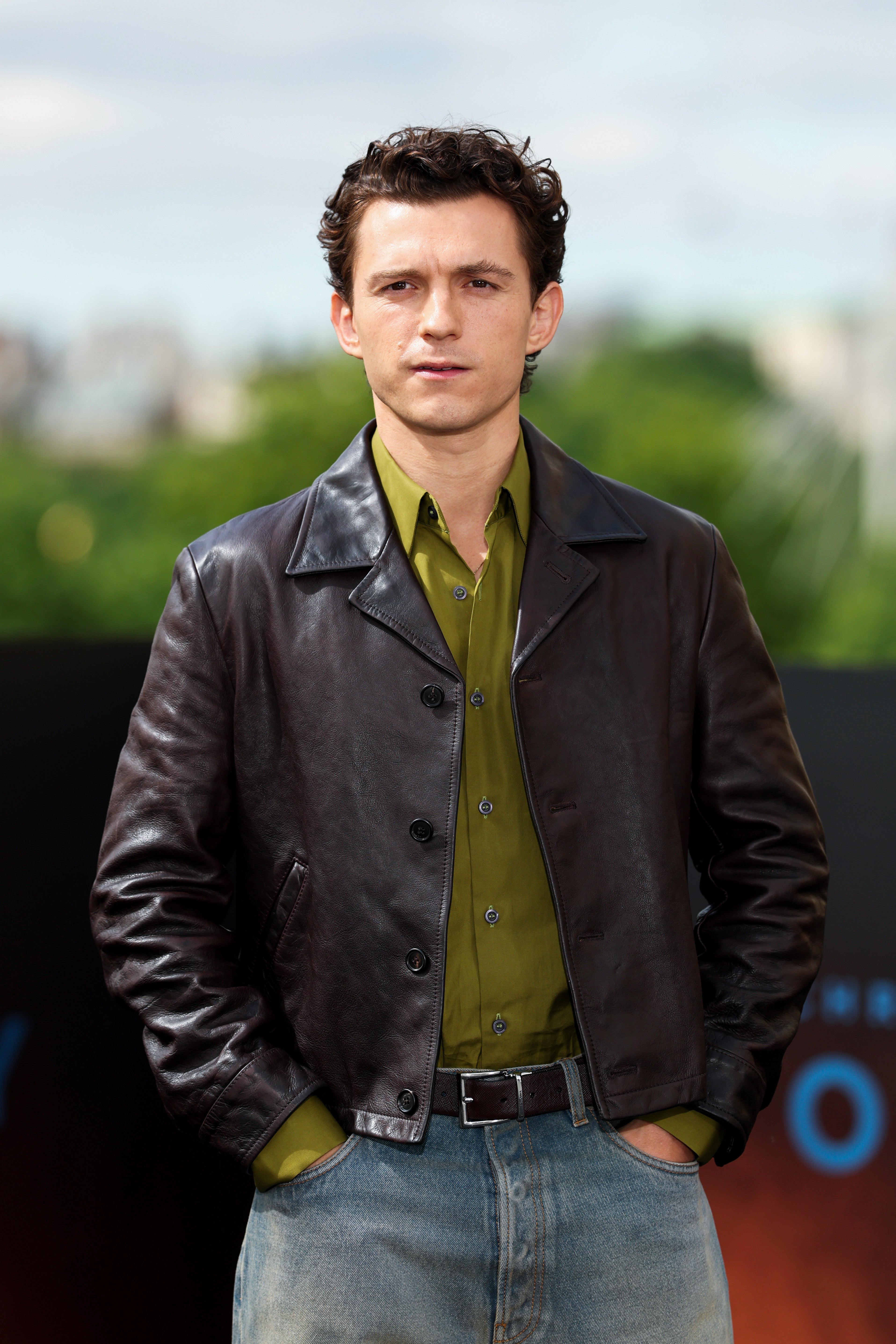Person in leather jacket and jeans standing outdoors, hands in pockets, at a public event