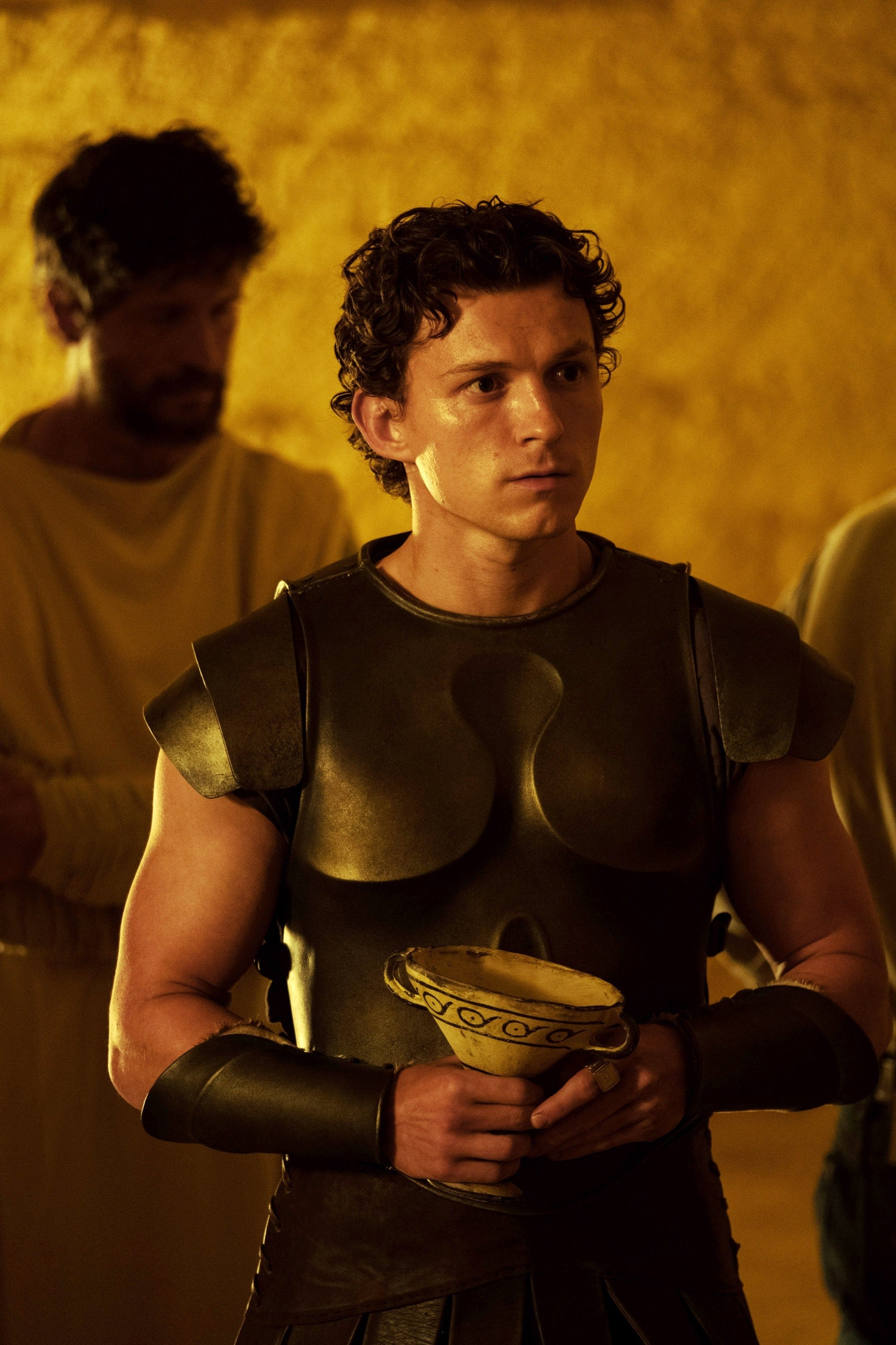 Person in period-style armor holds a chalice, standing in a dimly lit setting, with others in the background