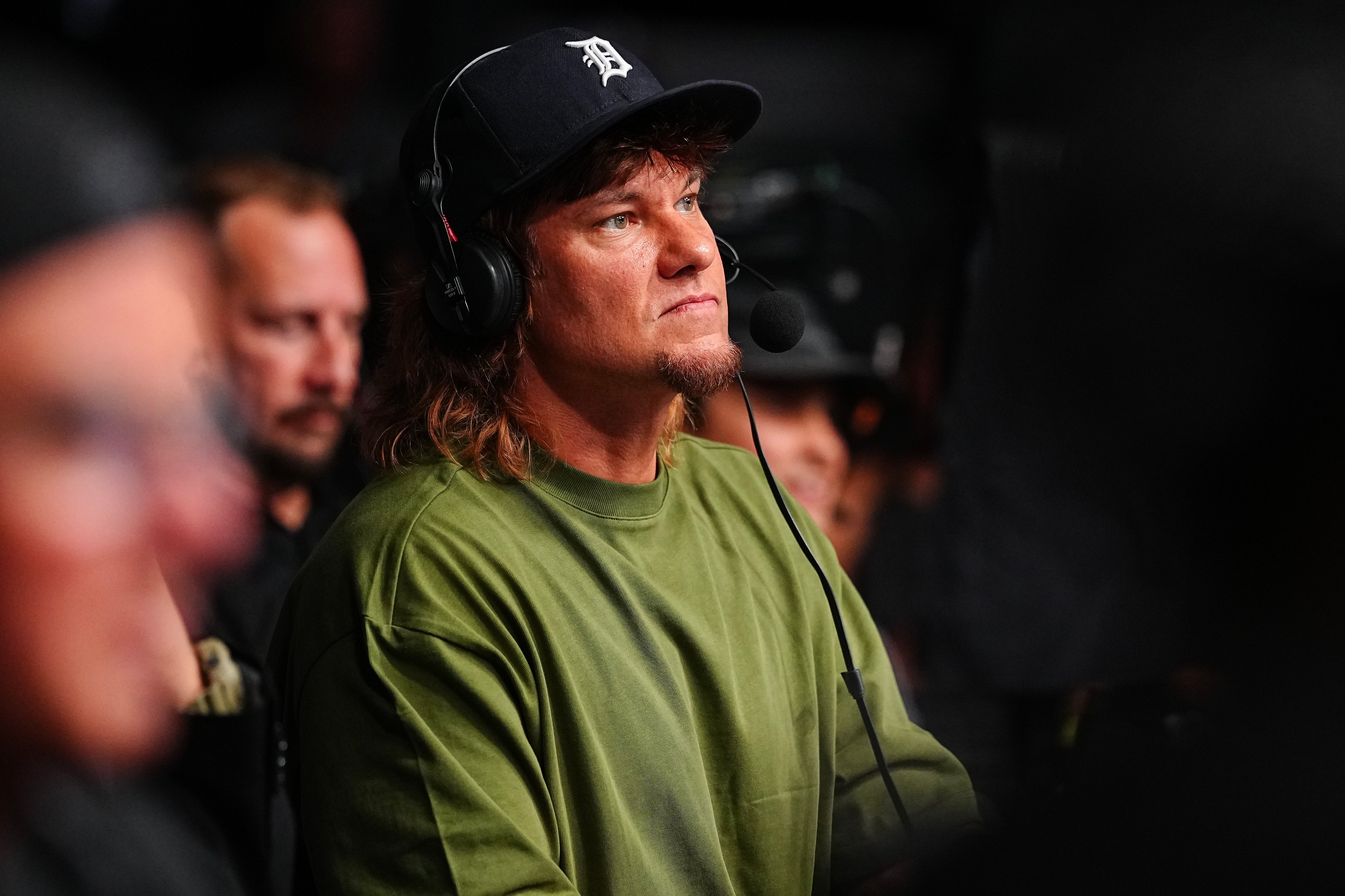Theo Von wearing a cap and headset, seated and focused, with others in the background, at a UFC  fight