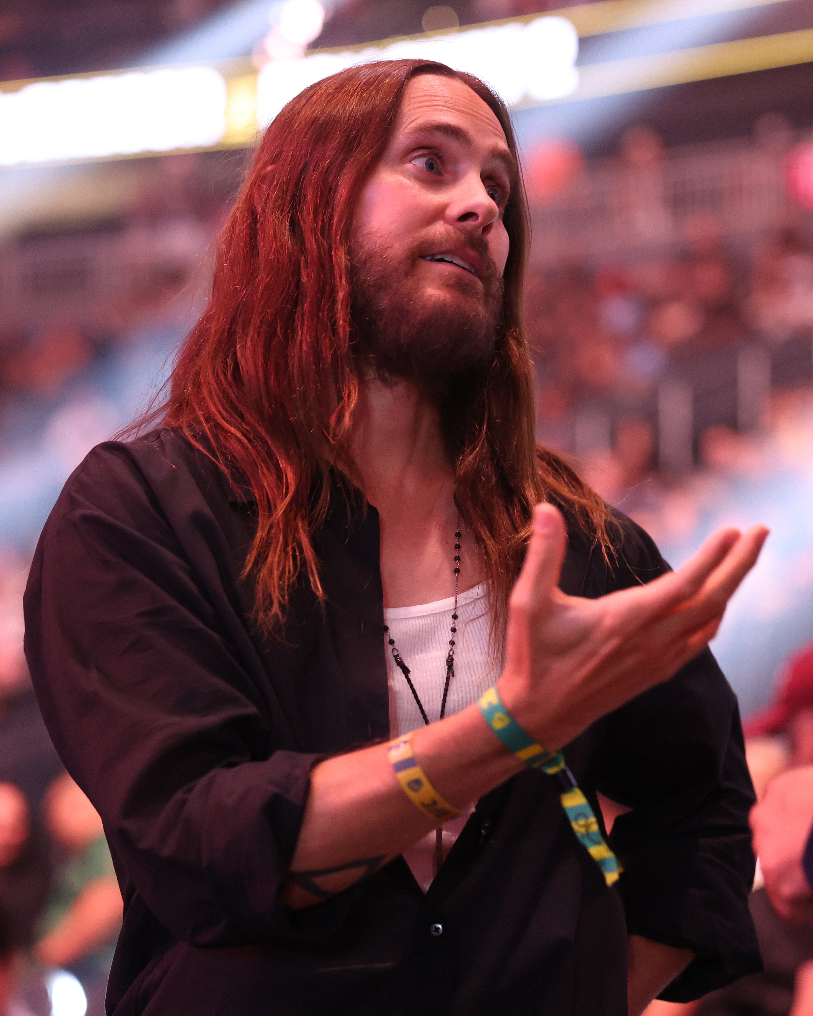 Jared Leto with long hair and beard in casual shirt, gesturing with one hand, wearing a festival wristband at a public event