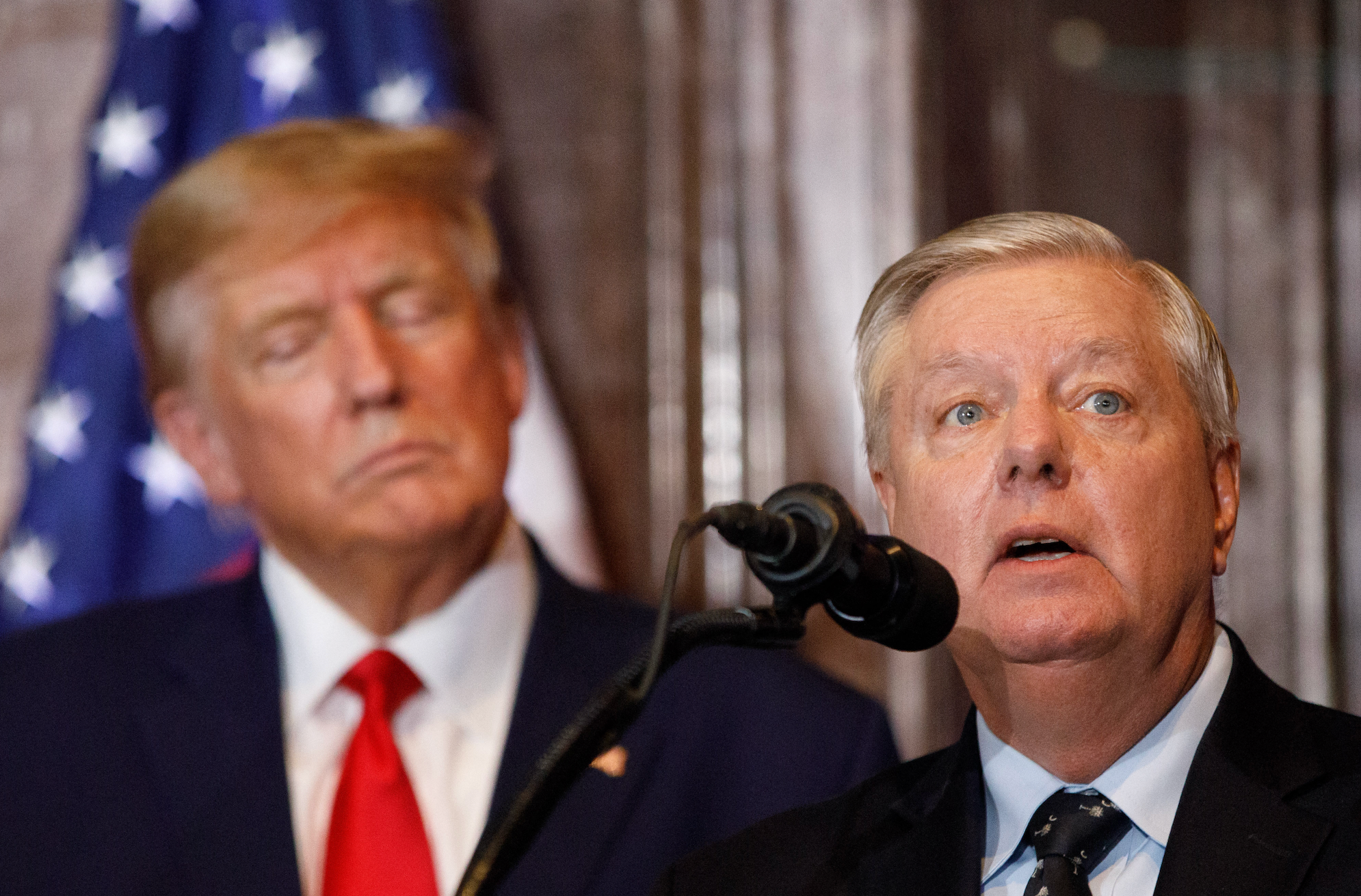 Donald Trump og Lindsey Graham på et arrangement; den ene på et podium med mikrofon, den andre lytter