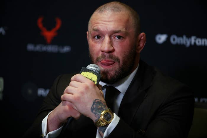 Conor McGregor in a suit at a press event holds a microphone, appearing thoughtful