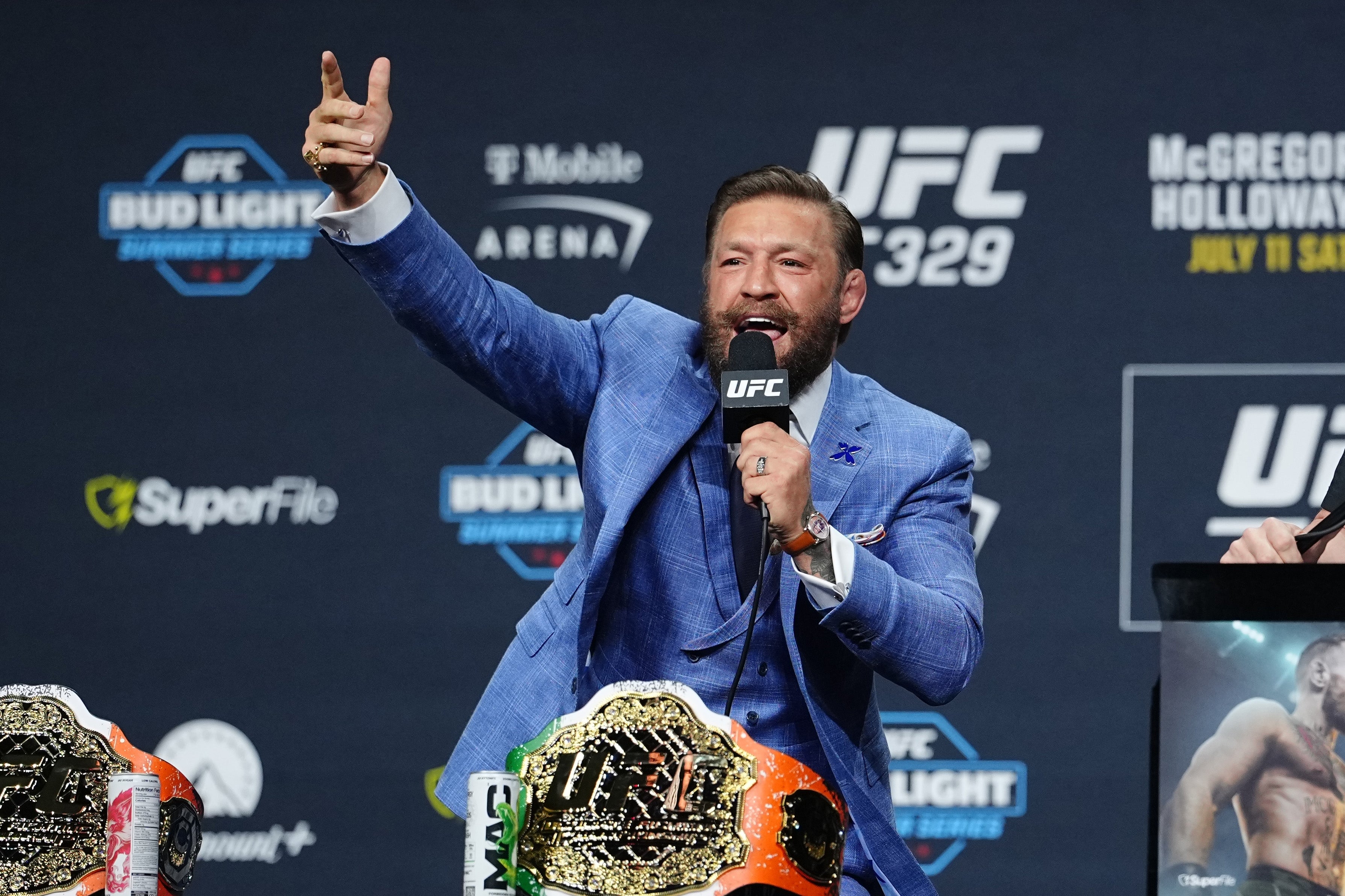 Conor McGregor in a suit energetically speaking at a UFC press conference, holding a microphone with championship belts displayed on the table