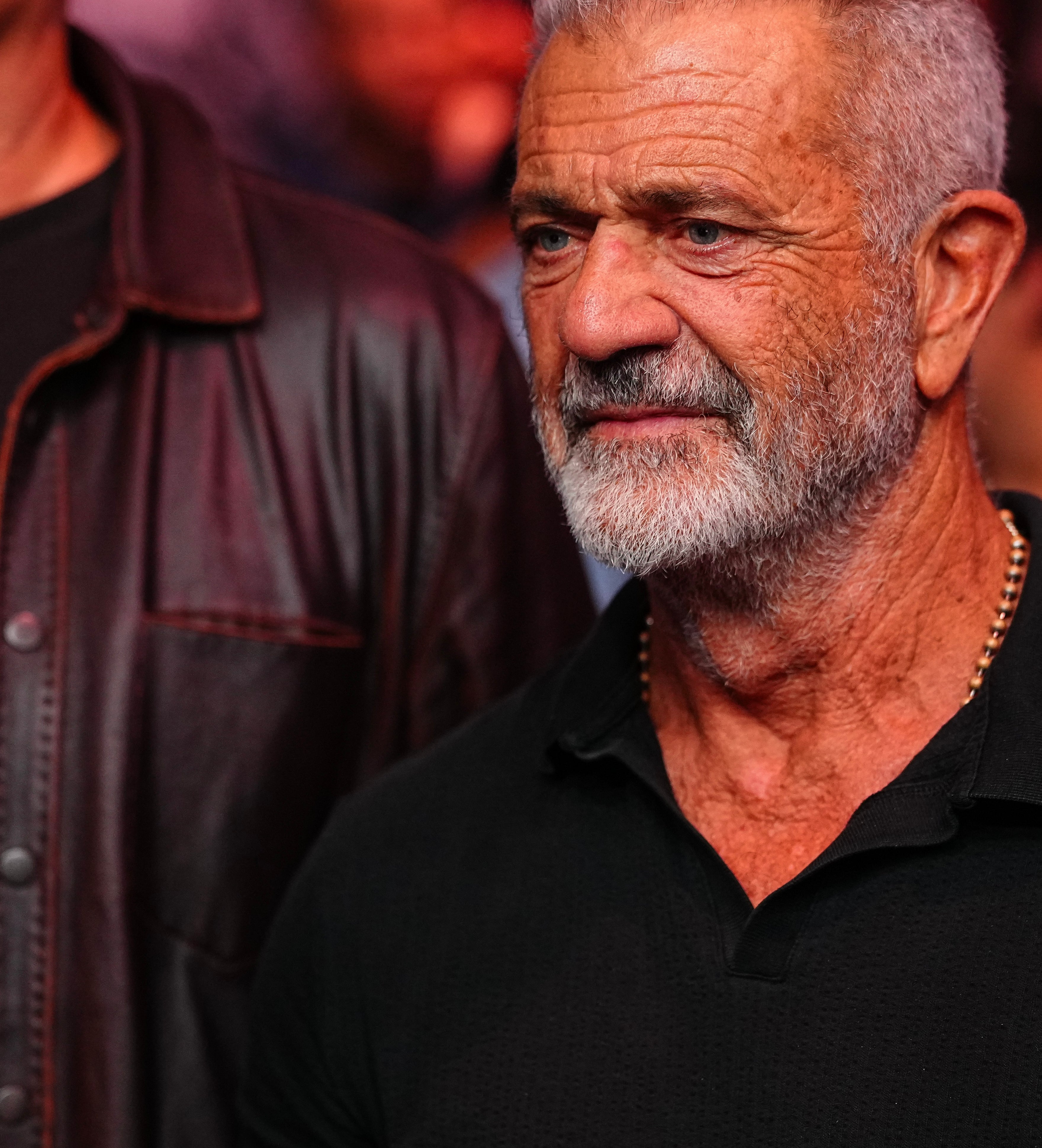 Mel Gibson in a casual polo shirt stands at an event, appearing engaged with the surroundings