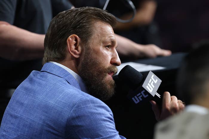 Conor McGregor in a suit speaking into a UFC microphone at an event