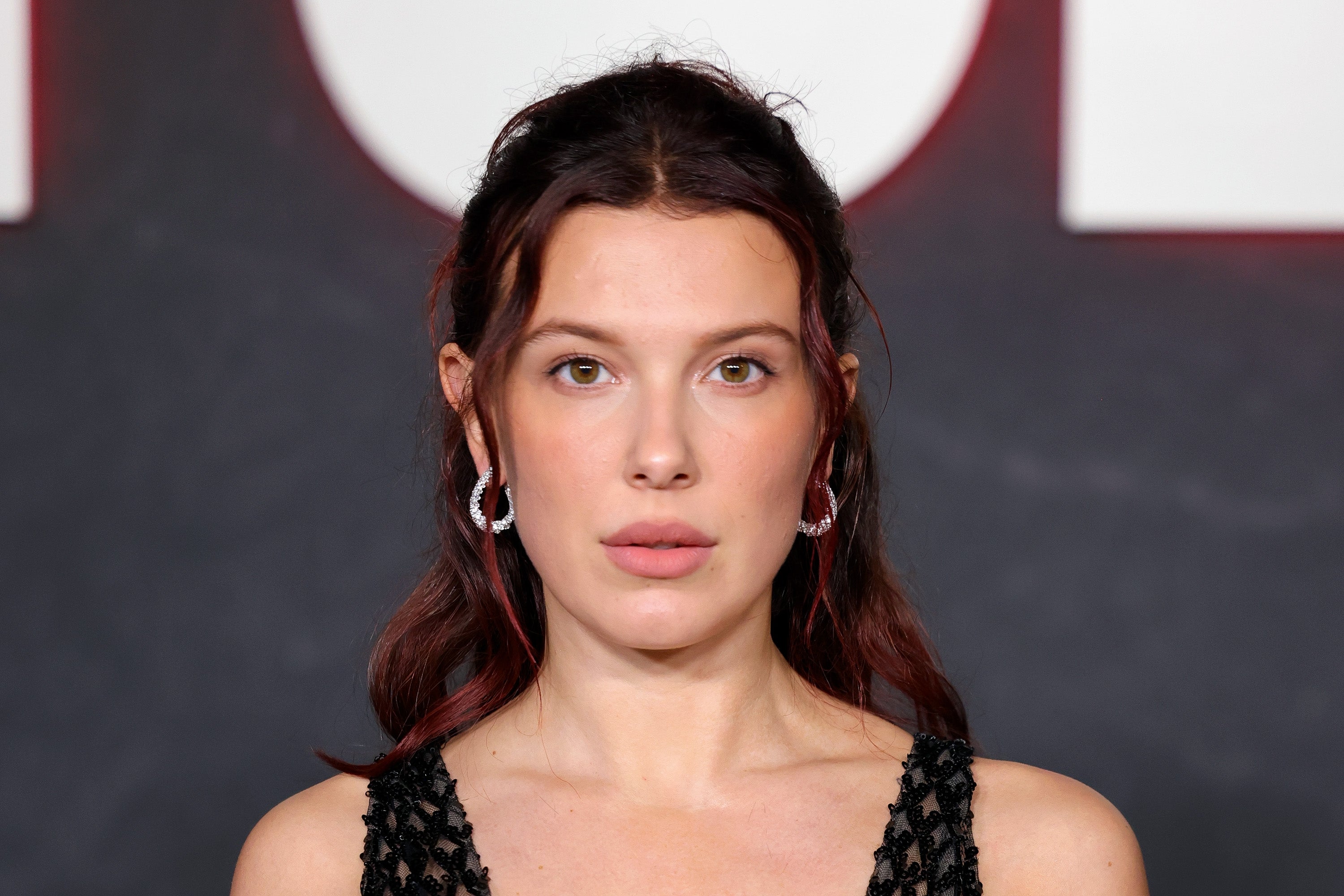 Millie Bobby Brown connected reddish carpet wearing a textured formal and earrings, posing successful beforehand of a backdrop