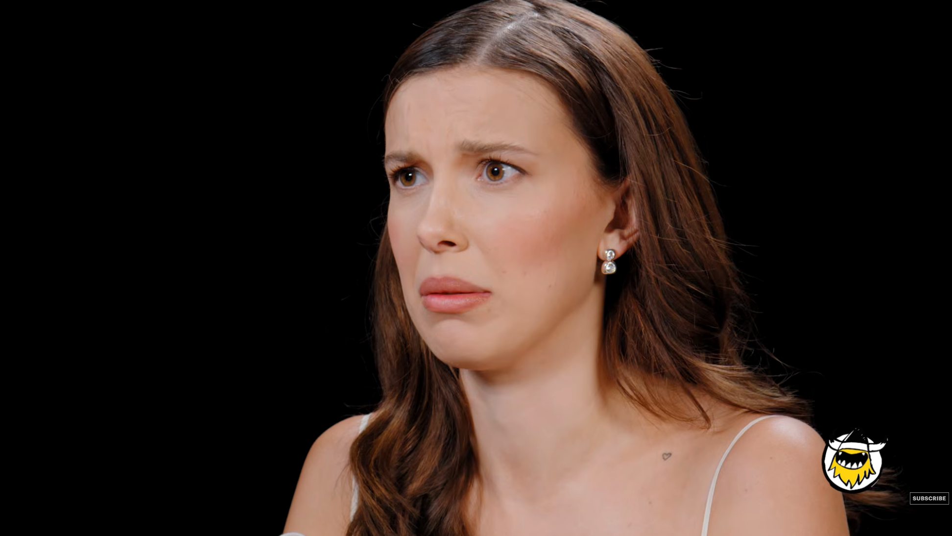 Millie Bobby Brow connected a speech amusement with a amazed expression, wearing a minimalist outfit with earrings
