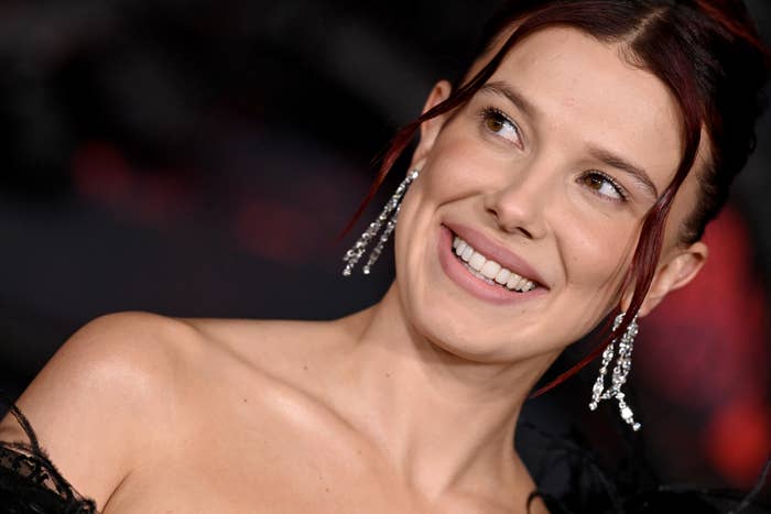 Millie Bobby Brown smiling, wearing elegant off-shoulder attire with sparkling earrings connected  a reddish  carpet