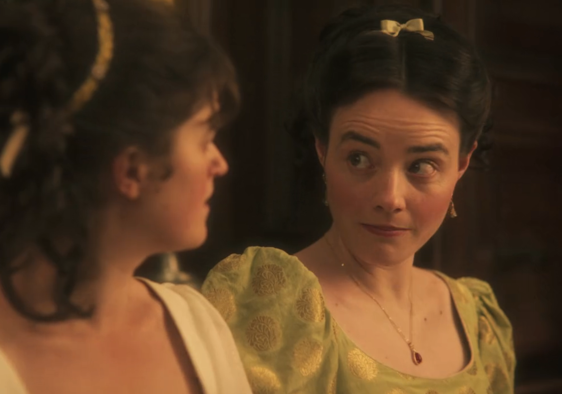 Two women in period attire share a moment, with one looking surprised or amused. They wear elegant, historical-style dresses with intricate hairdos