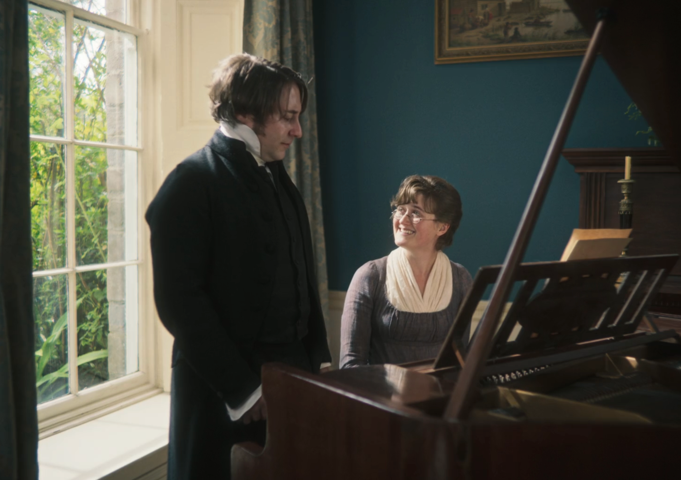 Two people in period clothing, one seated at a piano and the other standing, share a conversation in a sunlit room