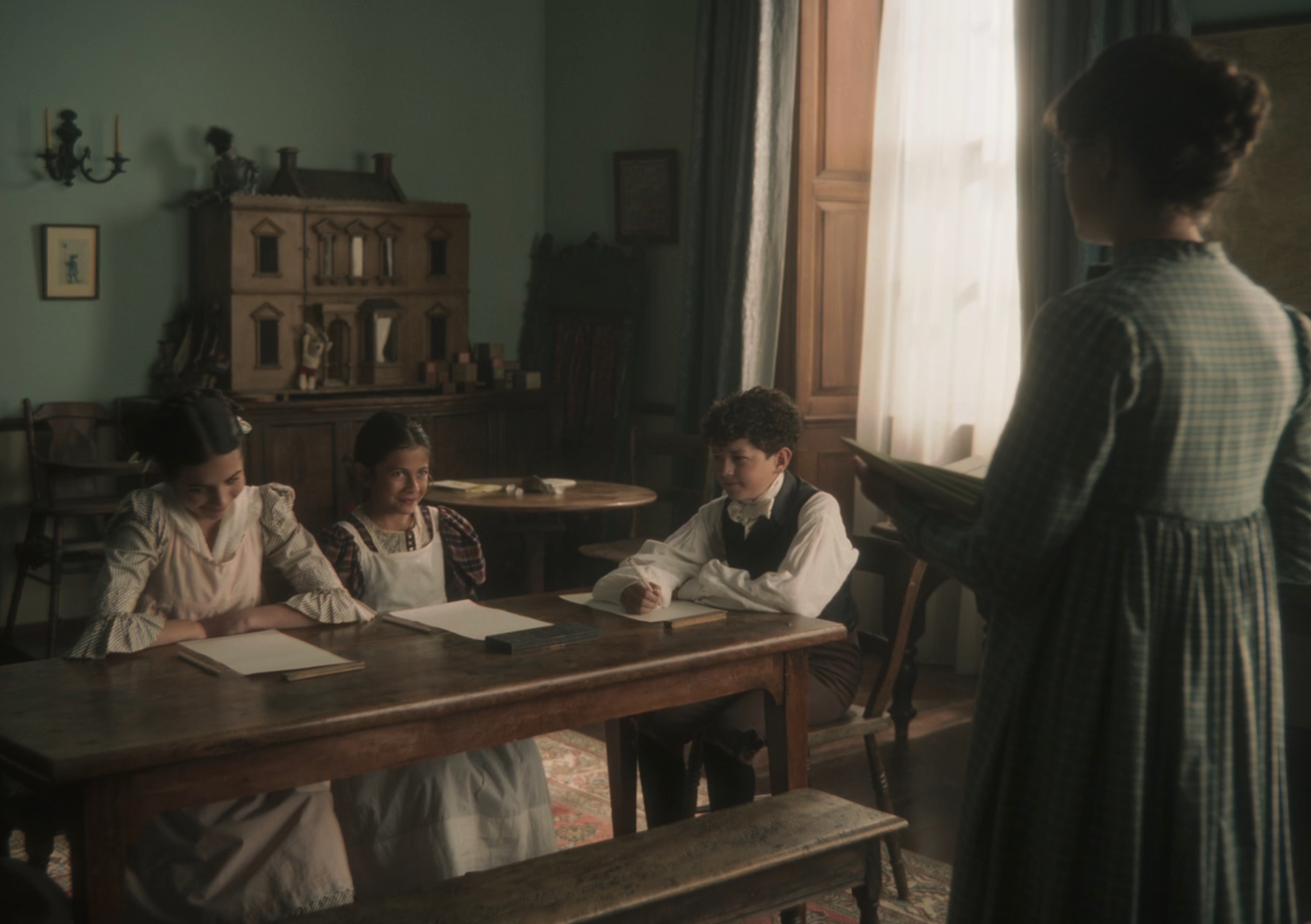 Three young students sit at a wooden table with papers, being instructed by a teacher in period clothing in a vintage classroom setting