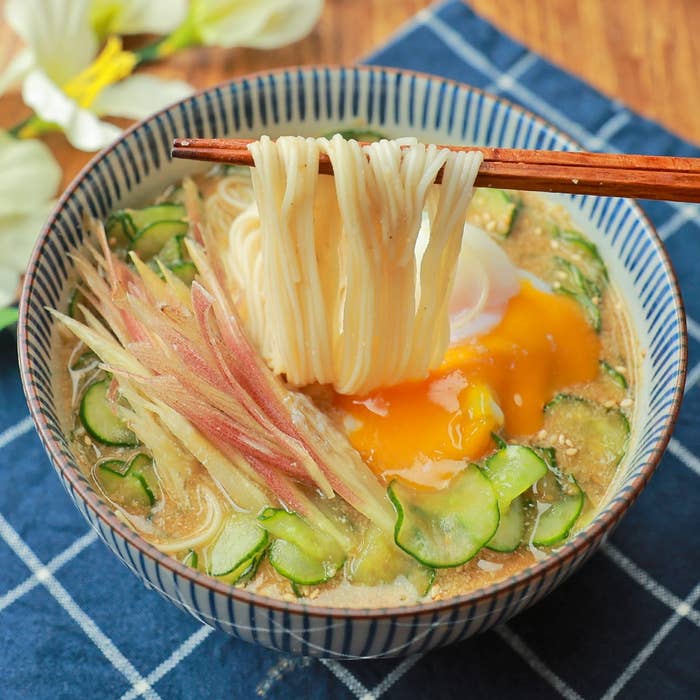 そうめんが箸で持ち上げられている。キュウリ、ミョウガ、卵もトッピングされ、美味しそうな一皿。